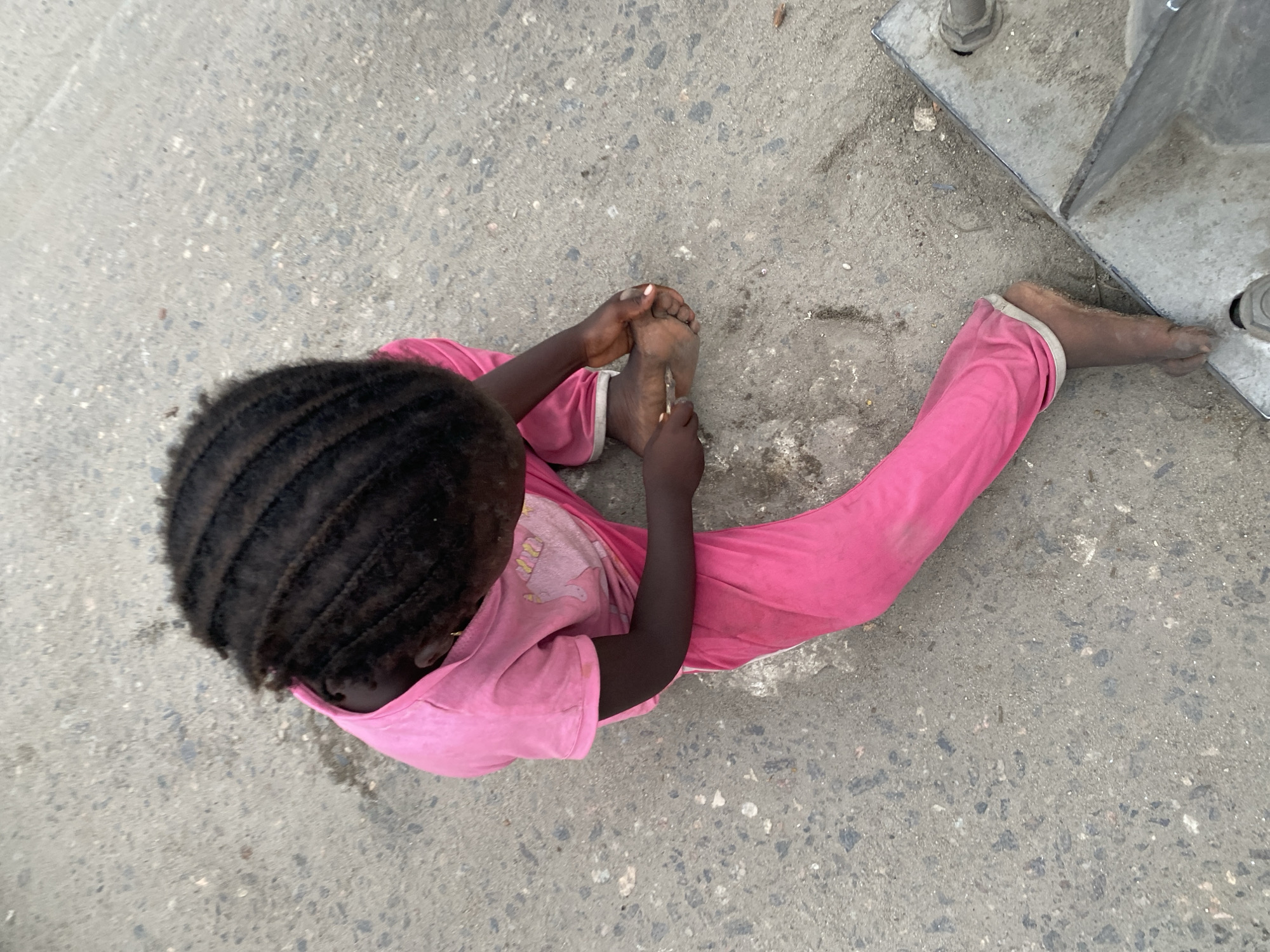 A child wearing pink clothing sits on a gray concrete surface. The child is examining the sole of their foot with a small object in their hand. The child's hair is braided. A concrete base of a structure, bolted to the ground, is partially visible on the right side of the image.