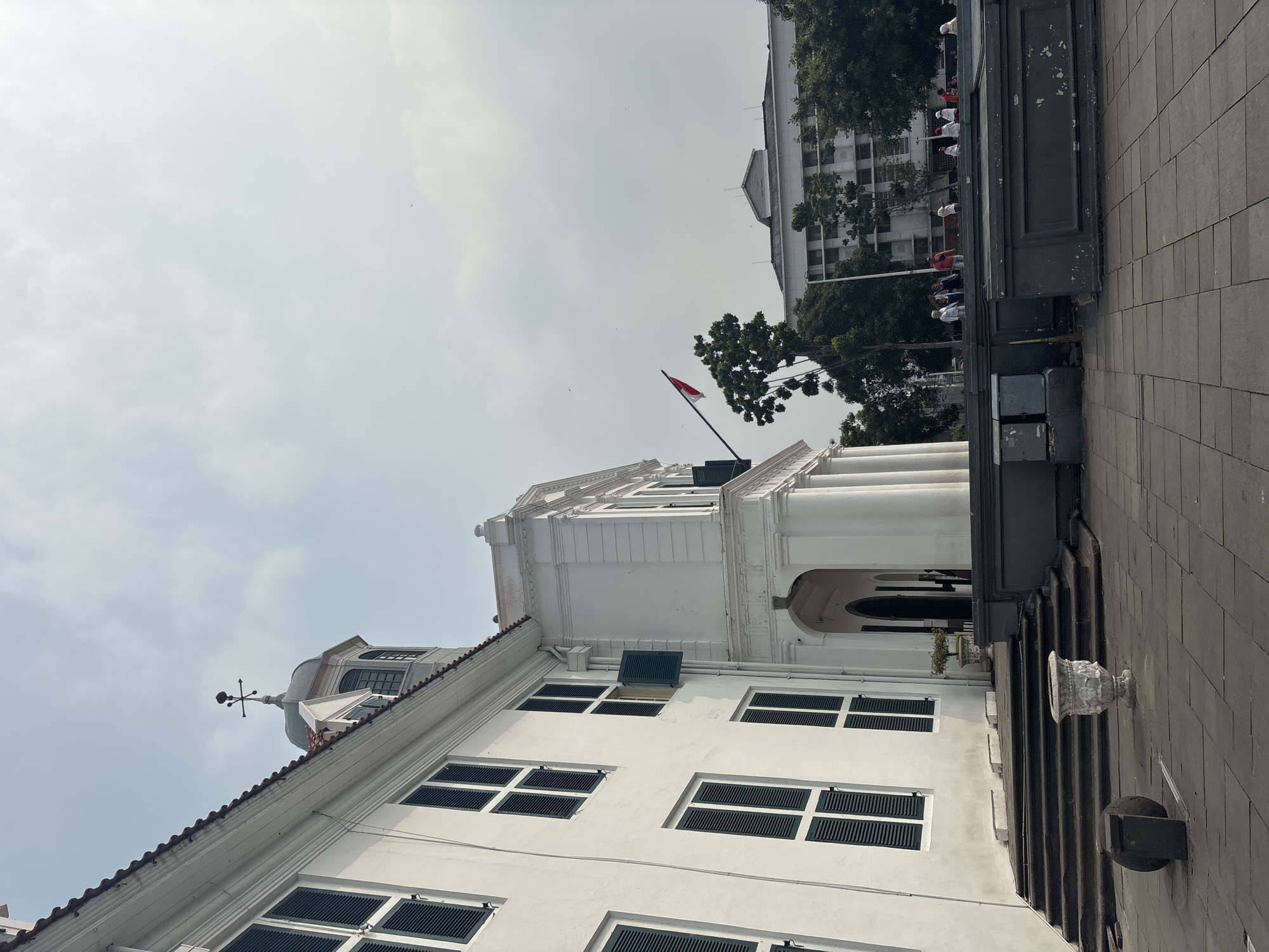 The image depicts the exterior of a white colonial-style building, likely a historical landmark in West Jakarta, Indonesia, given the architectural style and the Indonesian flag prominently displayed. The building features a row of dark-shuttered windows on its left side, and a portico with classical columns on the right. A dome with a weather vane crowns a section of the building. In front of the building, a paved courtyard is visible, with stone steps leading up to the entrance. A decorative urn and a stone sphere are placed in the foreground. Several people are seen in the distance, some walking and others standing, with a larger building and lush greenery in the background. The sky is overcast, suggesting a cloudy day. The scene captures a public space, possibly a museum or government building, given its grandeur and the presence of visitors.