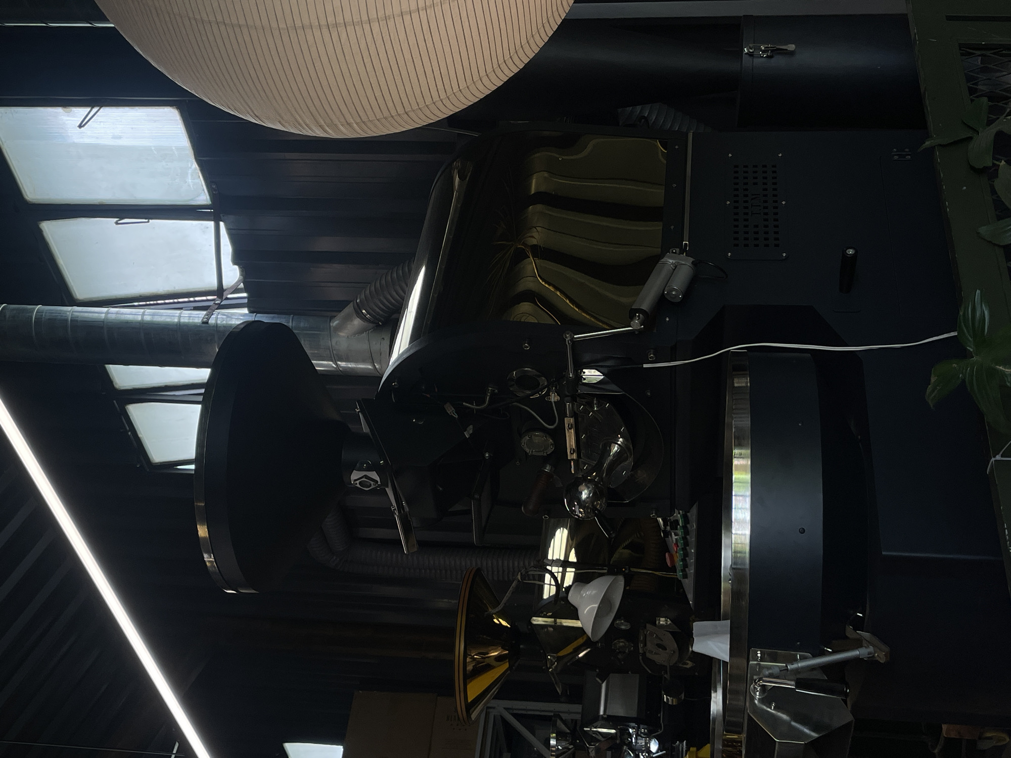 The image shows a complex piece of industrial equipment, possibly related to coffee roasting or processing, situated indoors. The scene includes a rounded, striped ceiling feature and overhead skylights suggesting a commercial or industrial space. The equipment itself is primarily black and metallic with various ducts, pipes, and electronic components visible. The location is Cam Ly - Đà Lạt, Vietnam, though no specific location details are provided other than the city.