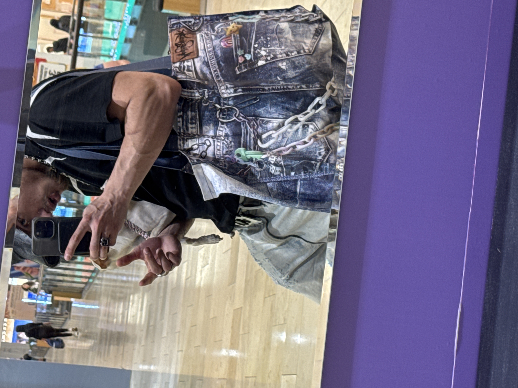 A mirror selfie shows a person with fair skin and dark hair, wearing a black t-shirt with white accents, and silver rings on their fingers. They are holding a smartphone up to take the picture. The reflection reveals a patterned denim tote bag. The background shows the interior of a building with light wood floors and display cases, possibly a shopping center. Outside the window is a reflection of passersby. There are no identifiable landmarks to confirm the location in Sofia, Bulgaria.