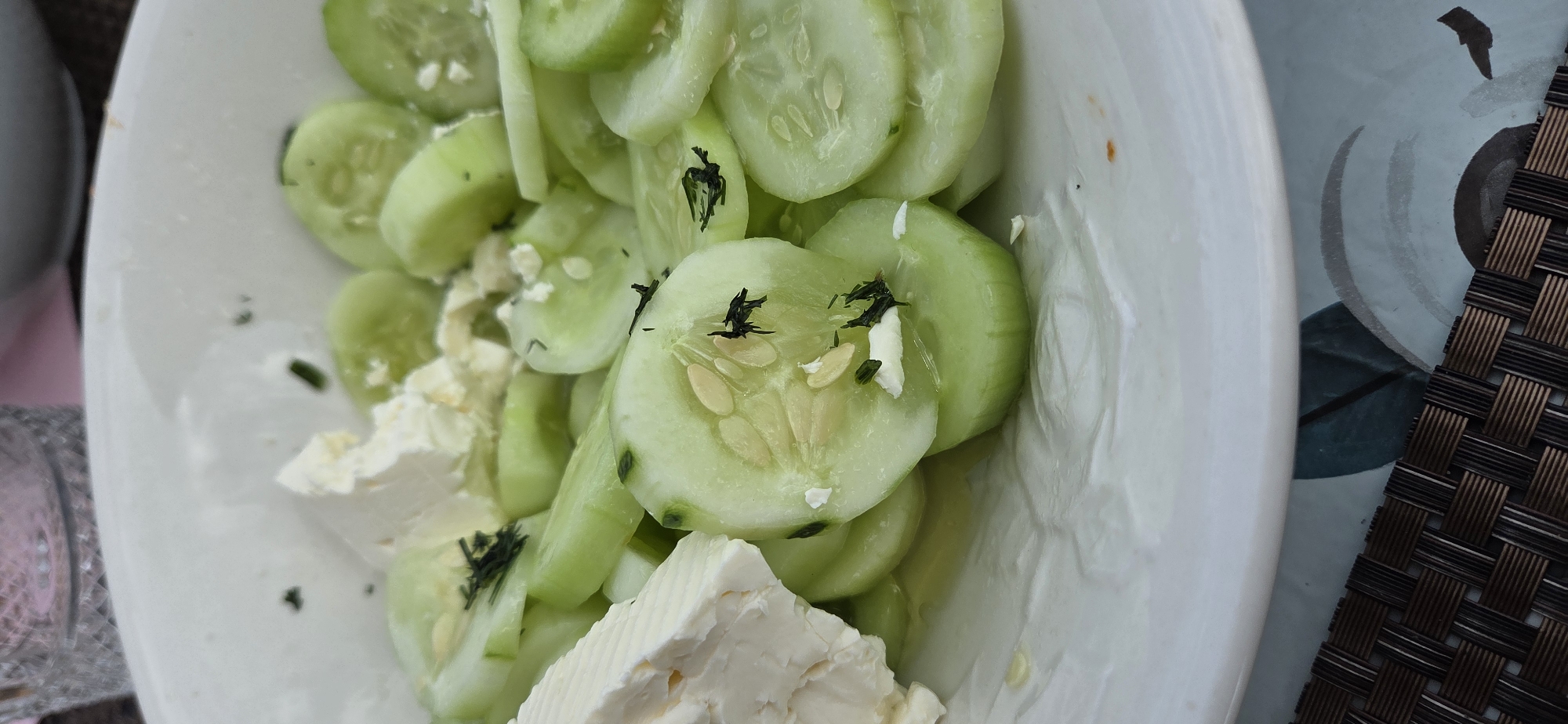 This image shows a close-up of a dish of sliced cucumbers and feta cheese, garnished with dill. The dish is served in a white bowl, and a portion of a patterned placemat and a glass are visible in the background. The lighting is natural, and the focus is on the food. The location is not identifiable as Eeklo, Belgium from the provided image.