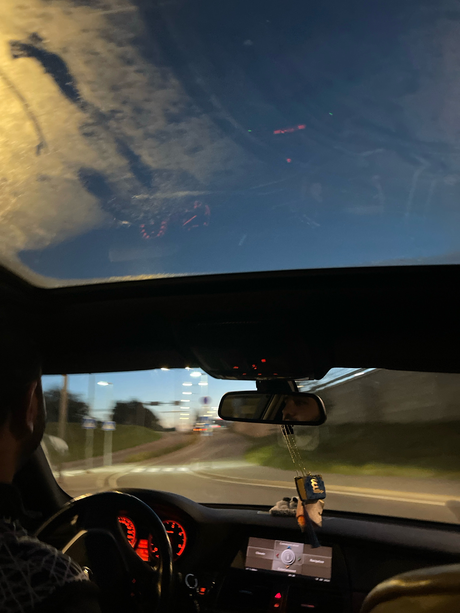 The image shows the interior of a car driving at night in Tallinn, Estonia, with a focus on the dashboard, steering wheel, and sunroof. The driver, visible in the rearview mirror, is wearing a checkered shirt. The car's dashboard lights are illuminated in red, and a car air freshener hangs from the rearview mirror. Outside the car, streetlights and traffic lights are visible, suggesting the car is on a road within the city. The sunroof offers a view of the dark sky, with reflections of what appear to be lights from the city and possibly clouds or other atmospheric phenomena. The overall scene captures a typical night drive in an urban environment.