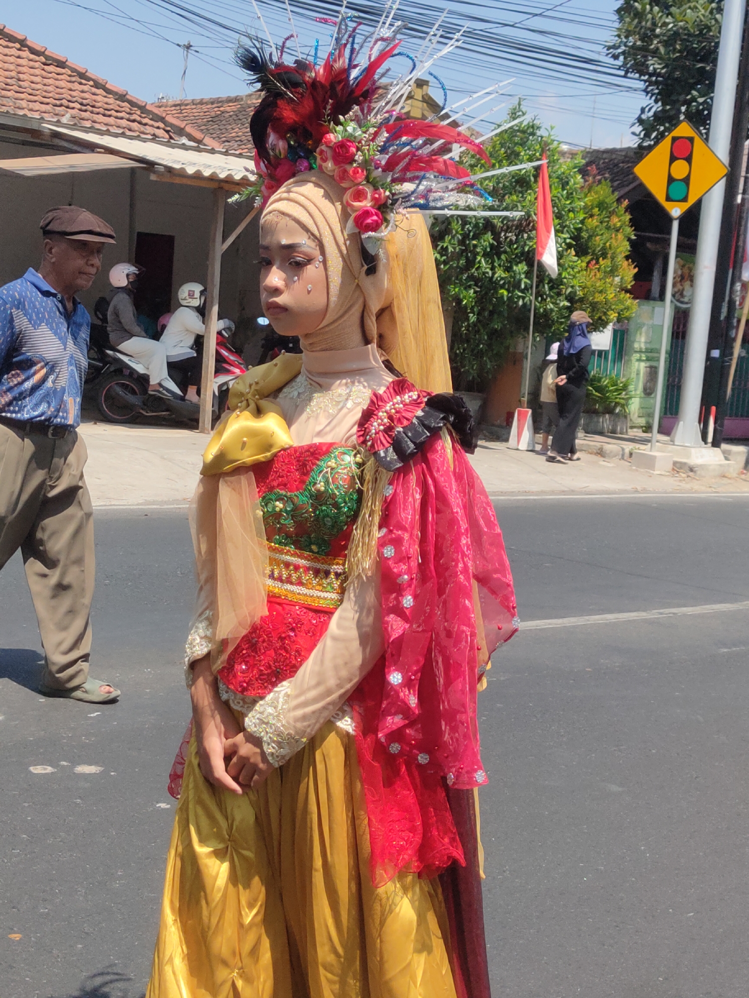 In the city of Ngasem, Indonesia, a young woman adorned in a striking, colorful costume stands on a paved road under a bright, sunny sky. Her attire includes a gold hijab, a vibrant red and green sequined bodice, and a flowing yellow skirt. She wears an elaborate headpiece featuring red feathers, flowers, and white spiky ornaments. Her face is decorated with white dots and lines around her eyes and cheeks. To her left, an older man wearing a blue patterned shirt and a brown cap walks away from the camera. In the background, buildings with red tile roofs line the street, and trees provide patches of shade. A red and white Indonesian flag flutters in the breeze, and a traffic light signal with yellow, red, and green circles is visible on a pole. Several motorcycles are parked along the side of the road, and other individuals are seen in the distance, some wearing headscarves. The overall scene depicts a cultural event or parade.