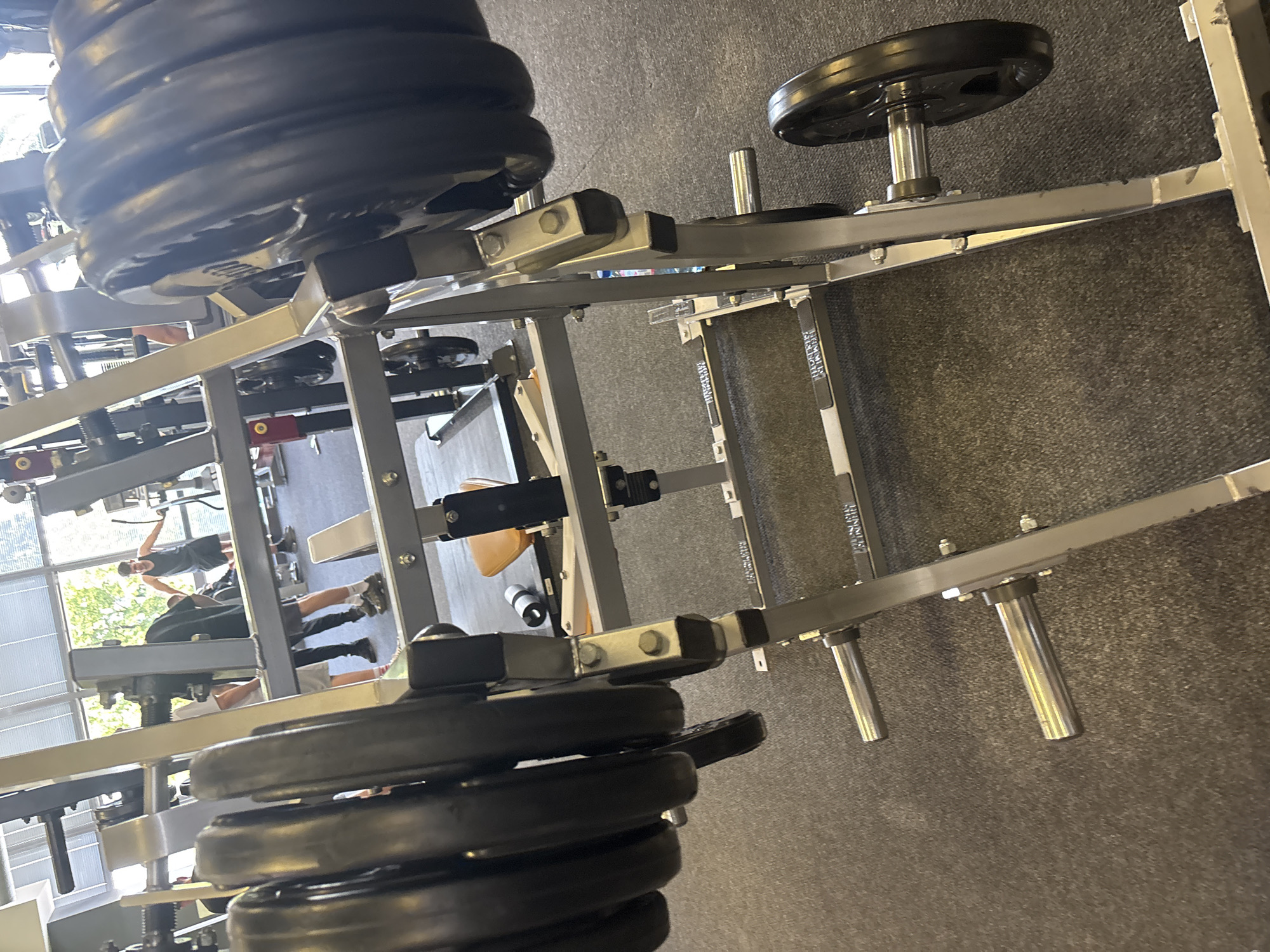 The image, taken in Blagoevgrad, Bulgaria, captures an indoor gym setting with multiple individuals and various pieces of exercise equipment. The perspective is from eye-level, looking across the gym floor towards large windows.
In the foreground, a substantial stack of black weight plates is visible, loaded onto a silver-colored, heavy-duty gym machine, possibly a plate-loaded leg press or similar equipment. The plates are round and uniformly black. The machine's frame is metallic, with visible bolts and sturdy construction.
The gym floor is covered with a dark grey, textured carpet. In the midground, other gym equipment can be seen, including parts of racks or benches. Additional weight plates are stored on a vertical peg extending from a silver frame on the right side of the image.
Through large windows in the background, bright natural daylight illuminates the scene, suggesting it is daytime. Green foliage from trees and the exterior of a building are visible outside the windows.
Several people are present in the gym, engaged in various activities. One individual, wearing dark shorts and a dark top, stands near the windows with arms seemingly raised or stretching. Another person, partially obscured by equipment, wears a white t-shirt and dark shorts. Legs of a third person, in dark trousers and white socks, are also visible behind some machinery.
Visible text includes the brand "HAMMER STRENGTH" on a black strap or part of a machine in the midground, and possibly on a white band near the floor. The overall atmosphere appears active and functional, typical of a commercial or institutional fitness facility.