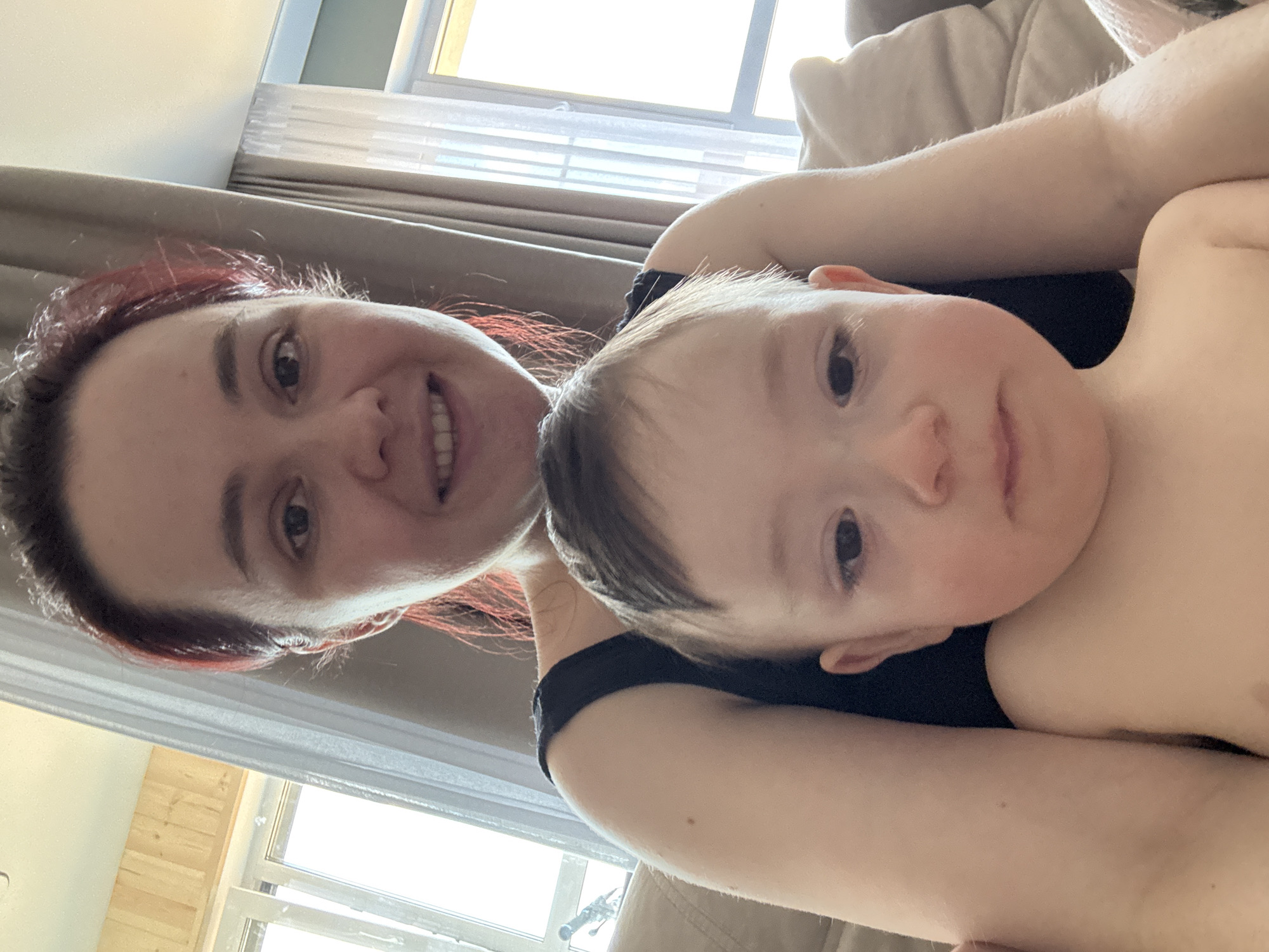 A selfie shot shows a woman with auburn hair smiling at the camera while holding a baby in her arms. The background includes a window with curtains and what appears to be a couch. There is no information available to identify the location as Izhevsk, Russia.