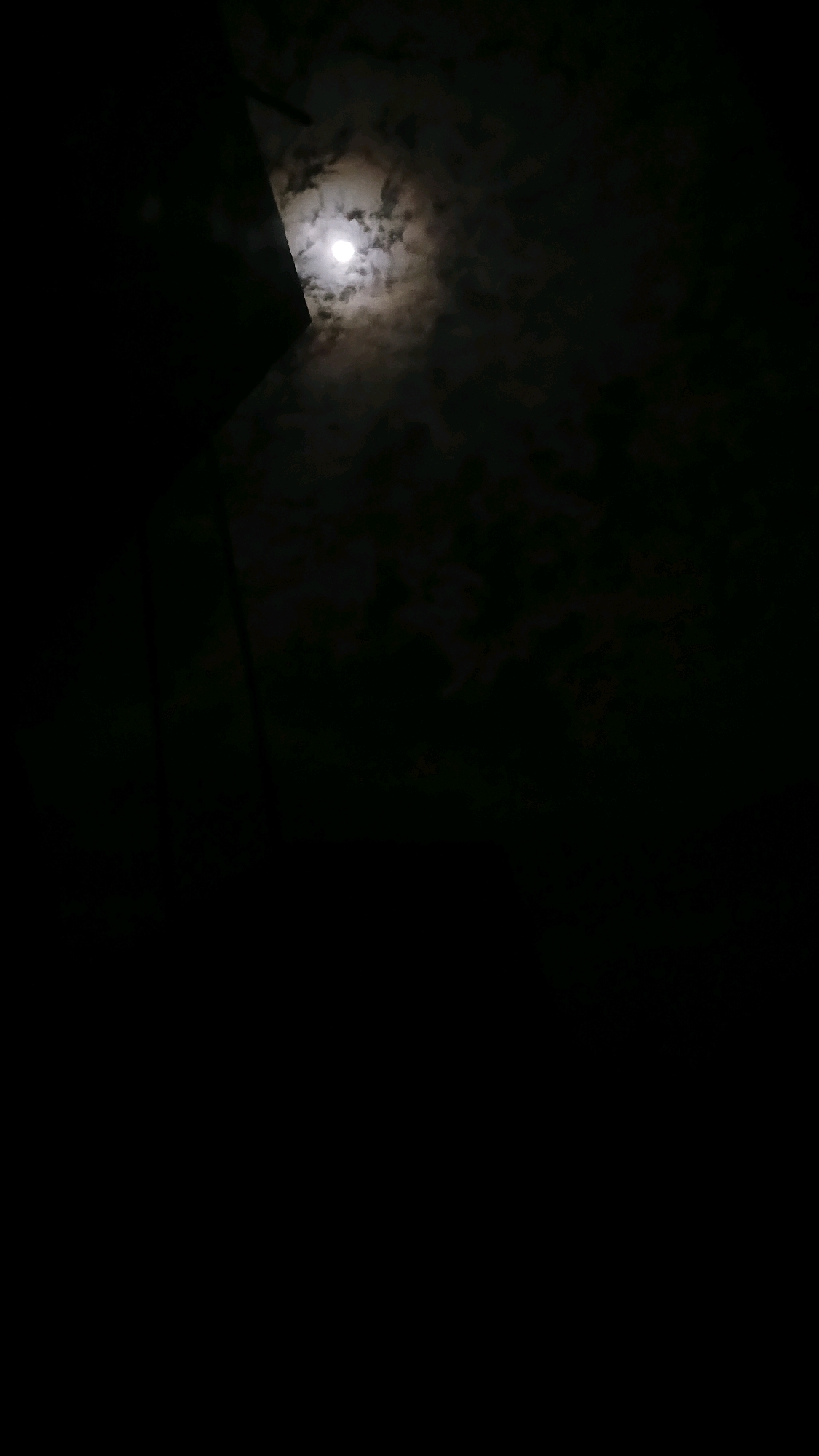 The image shows a dark night sky with a bright full moon peeking through scattered clouds. To the left, a dark, angled structure, possibly a building or part of a roof, partially obscures the moon. The majority of the image is black, indicating deep shadow or nighttime darkness. There are no people or discernible actions visible. The scene appears to be an outdoor setting, likely urban given the presence of the structure. Without further information or context, the specific location in Haivoron, Ukraine cannot be determined from the visual elements alone.