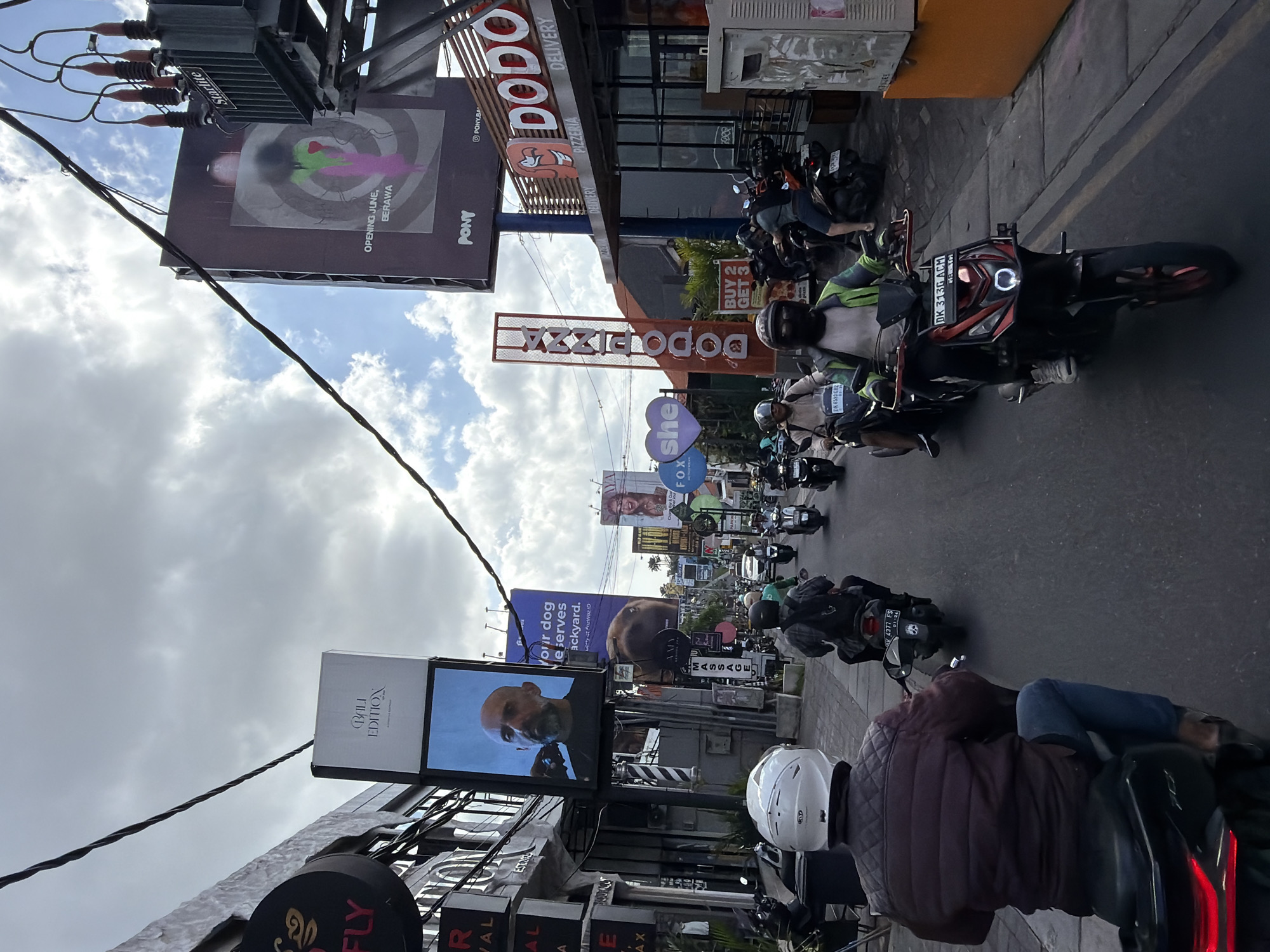 Captured from the vantage point of a motorbike rider, the image presents a bustling street in North Kuta, Indonesia, lined with establishments and assorted advertisements. In the foreground, other motorbike riders navigate through the street. Prominent establishments include Bali Edition and Dodo Pizza, the latter identified by several signs. Street-level advertisements include those for veterinary services and massage parlors, alongside larger commercial displays. Overhead, additional advertisements for Pony are visible. The urban environment is framed by a sky marked with scattered clouds.