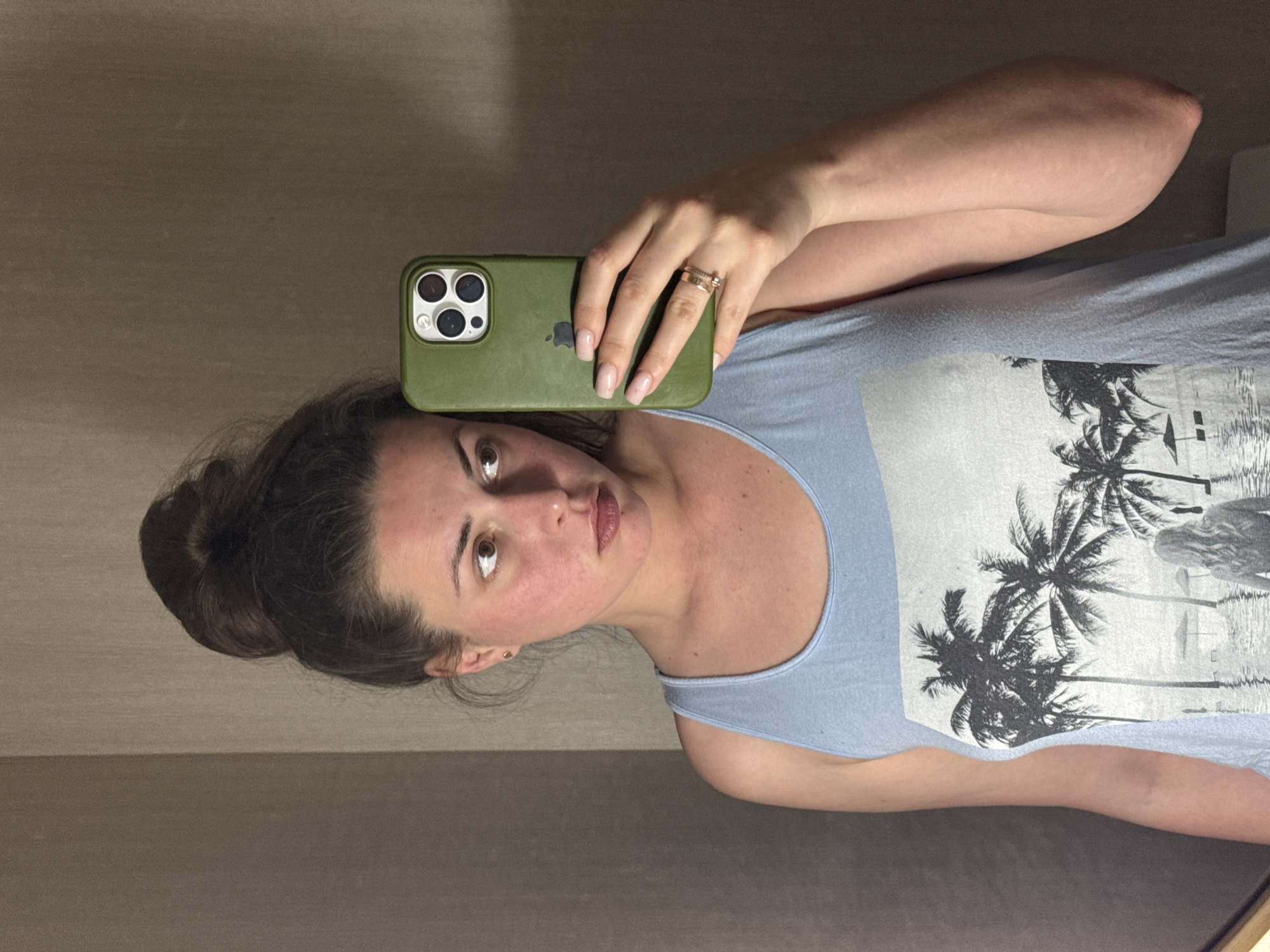 A woman is taking a selfie in a mirror. She is wearing a light blue tank top with a palm tree graphic. Her hair is pulled up in a messy bun. She is holding a green phone in her right hand, and a ring is visible on her ring finger. The background appears to be a room with textured walls. The lighting is dim, and the overall scene suggests an indoor setting, possibly a hotel room or a bathroom. There are no visible indications of a specific city or location.