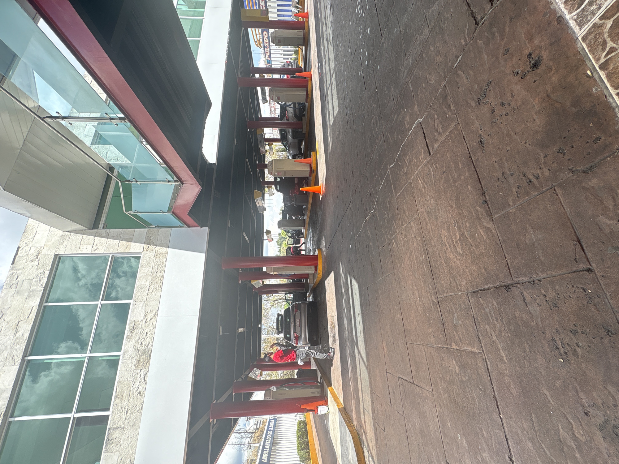 The image captures a view of a car wash establishment in Huixquilucan de Degollado, Mexico. The establishment is characterized by red support pillars beneath a covered area, sheltering a row of car washing stations. Vehicles are parked at some of the stations. An individual is seen near one of the cars wearing a red shirt. Orange traffic cones are placed at intervals near the car wash stations. A multi-story building with glass windows and a stone facade adjoins the car wash area. The foreground is occupied by a paved walkway with a textured stone surface.