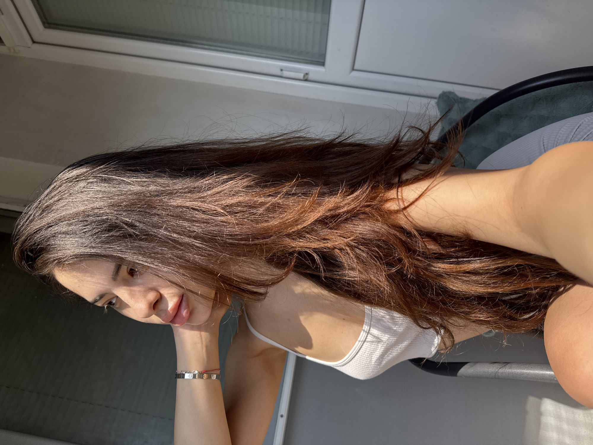 A young adult female is depicted in a brightly lit indoor or balcony setting in Plovdiv, Bulgaria, captured in a self-portrait. She is shown in profile, looking towards the left side of the frame with a pensive expression. Her long, wavy dark brown hair with reddish highlights flows over her shoulders. She wears a white ribbed camisole and a silver-toned bracelet featuring interlocking motifs, accompanied by a thin red string, on her left wrist. Her left arm is raised, with her hand resting at the back of her neck/head. Strong, direct sunlight from the right casts harsh shadows across her face, arm, and chest. She appears to be seated on a chair with a dark metal frame and a visible teal, textured cushion. The background on the left features horizontal slat blinds or shutters, while the right side displays a white-framed glass door or window, also with interior horizontal blinds, suggesting an exterior view.