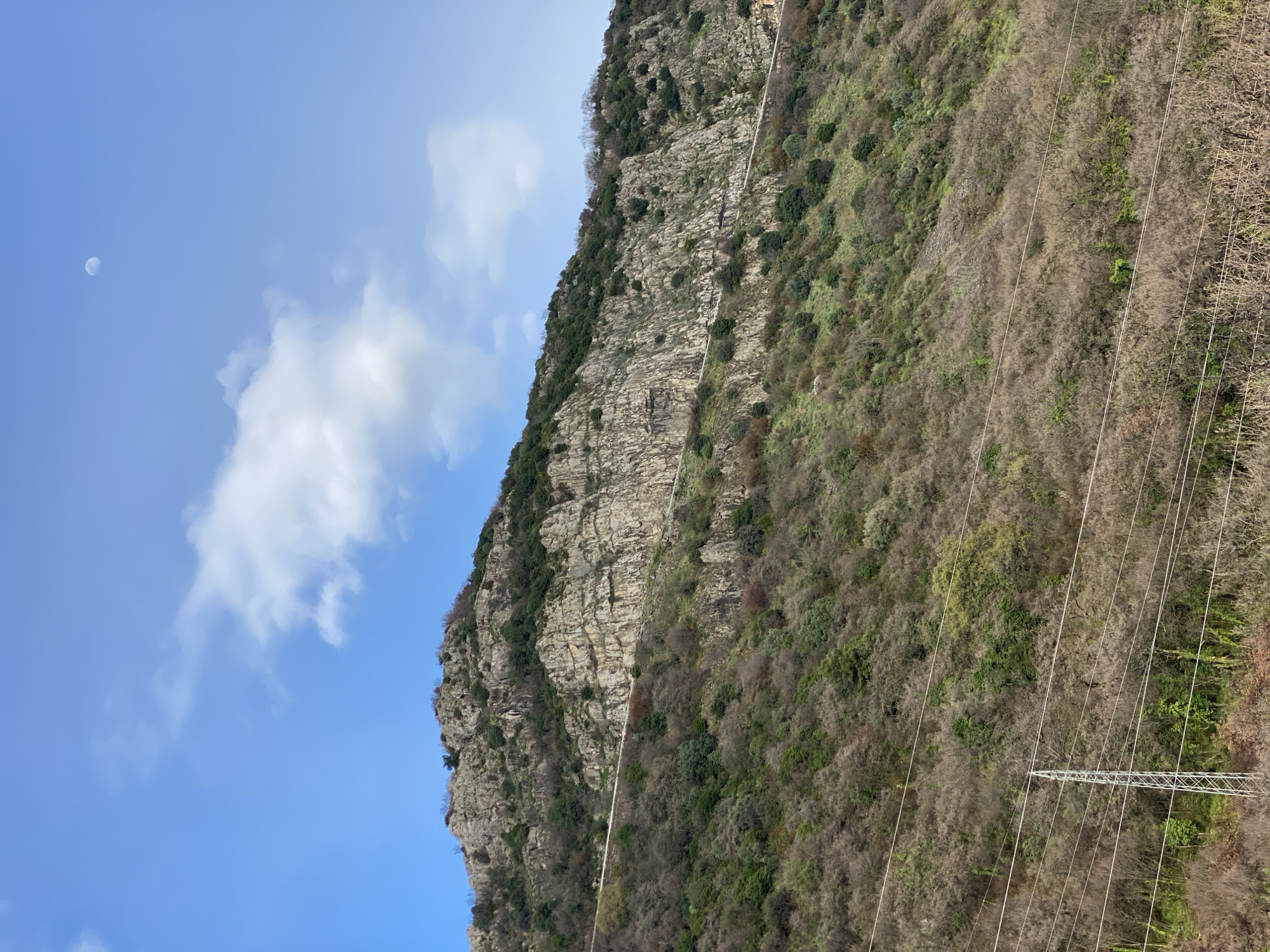 The image displays a steep, rocky mountainside in the vicinity of Budva, Montenegro, under a clear blue sky with scattered white cumulus clouds. A distinct crescent moon is visible in the upper right portion of the sky. The upper and middle sections of the mountain feature exposed grey rock formations and cliffs, interspersed with dark green evergreen foliage. A narrow, light-colored, horizontal road or path is discernible traversing the mid-mountain slope. The lower two-thirds of the mountain are densely covered with vegetation, comprising a mix of evergreen shrubs and numerous bare deciduous trees, suggesting a cool season. In the lower left foreground, a partial view of a metal lattice power transmission tower is present, with multiple parallel power lines stretching horizontally across the lower frame. No individuals or active subjects are depicted.