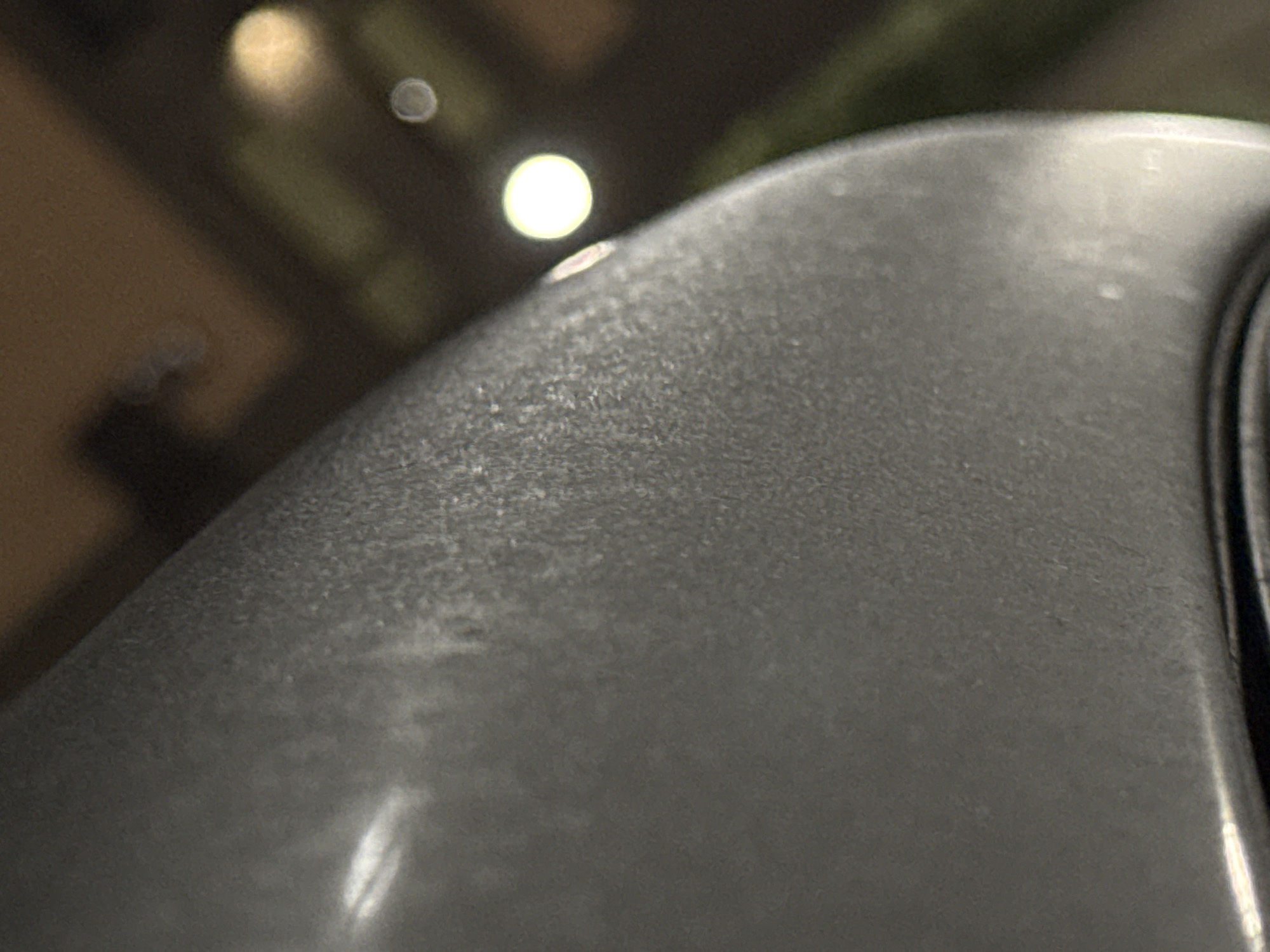 The image shows a close-up, low-angle shot of a textured gray surface, possibly a vehicle or outdoor structure. The surface has a matte finish with subtle variations in tone and texture. In the background, out of focus, are hints of an urban environment at night. There are bright, circular lights, suggesting streetlights or building lights. The ground appears to be paved, with some green foliage visible on the right side. The overall impression is of an outdoor scene in a city, with the primary focus on the detailed texture of the foreground object. There is no specific information that places this image in Markova, Russia.