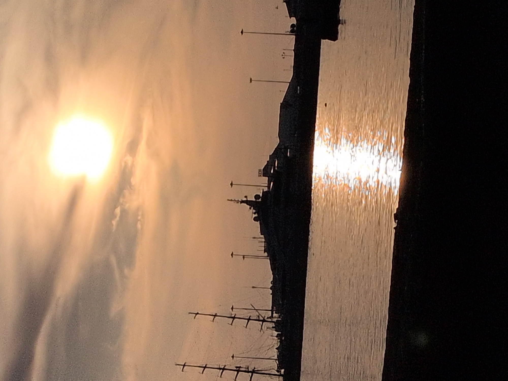 The image depicts a coastal or harbor scene at either sunrise or sunset, characterized by strong backlighting creating silhouettes. The sky, occupying the upper two-thirds of the frame, displays warm hues of orange, yellow, and muted grey. The sun, appearing as a bright, overexposed white disc, is positioned in the upper center-right, casting a prominent, shimmering reflection on the dark, calm water below. Scattered, thin clouds are visible in the sky.
In the mid-ground, the dark silhouettes of several large vessels and shoreline infrastructure are present. On the left, a multi-masted sailing yacht or tall ship is visible, its rigging and masts clearly silhouetted against the sky. To its right, a large motor yacht or ship, with a distinctive superstructure featuring multiple decks and antennae, is also silhouetted. Behind and between these vessels, a dark, elevated landmass or extensive dock structure runs horizontally, punctuated by numerous tall, slender poles that are likely streetlights or dock lights. A small, lighter-colored detail, possibly a sign with the letters "KCH" or similar, is faintly discernible on the landmass or ship's superstructure in the central mid-ground.
The immediate foreground, occupying the bottom approximately 15-20% of the image, consists of a dark, undifferentiated landmass or quay. No individuals are discernible in the scene. The location is the city of Sochi, Russia.