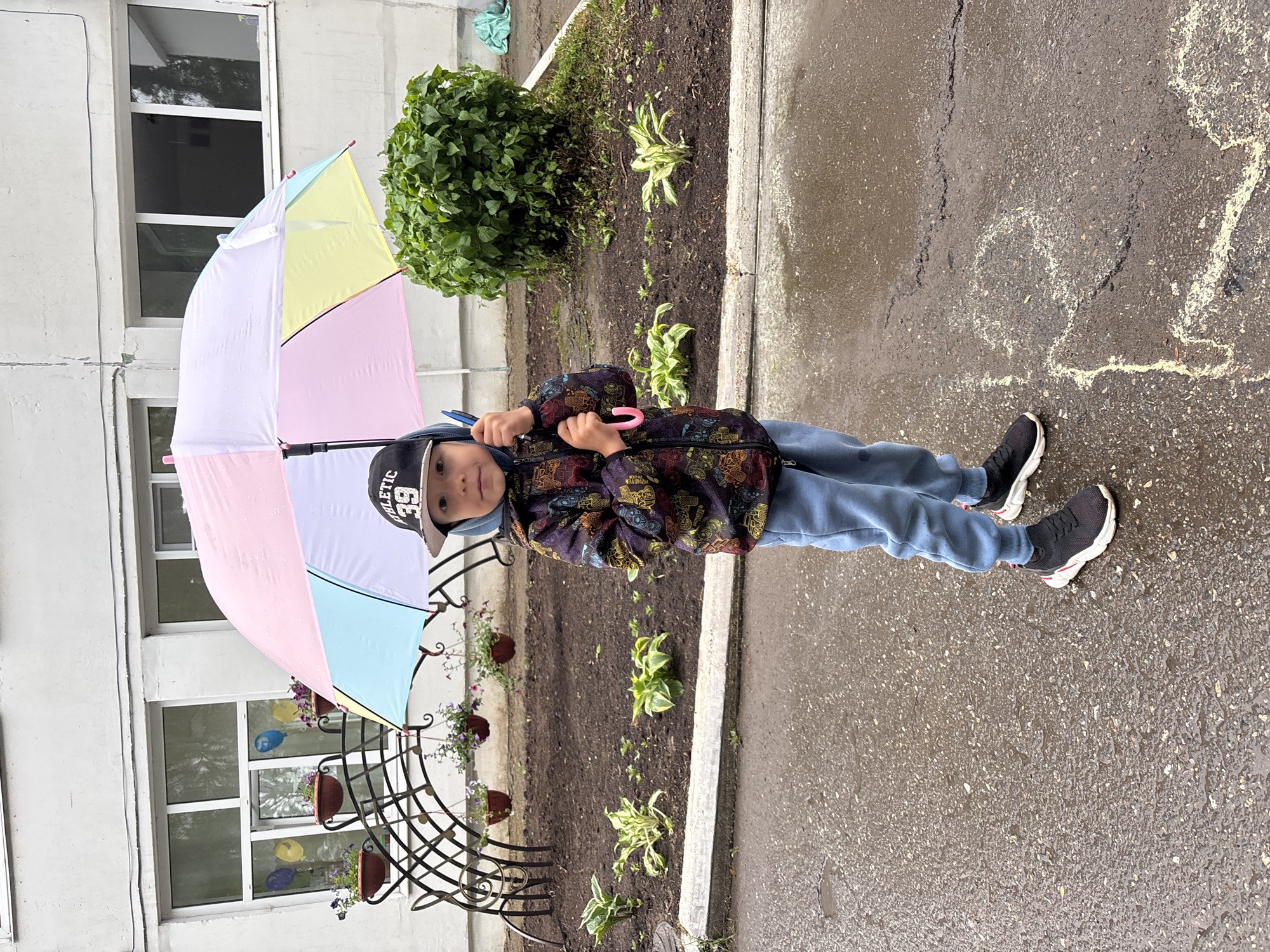 A child stands on a wet pavement surface, holding a pastel-colored umbrella. The child is wearing a cap, a patterned jacket, and blue pants. To the left is a building with a metal structure holding flower pots. Behind the child is a flower bed with a bush. A drawing is visible on the pavement. The image was taken in the city of Chaikovsky, Russia.