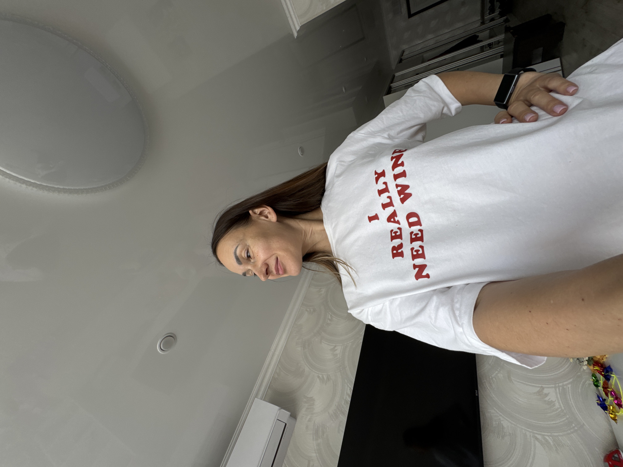A woman with shoulder-length brown hair, wearing a white t-shirt with red lettering that reads "I REALLY NEED WINE", stands in what appears to be an indoor residential setting. The t-shirt has a loose fit. She is looking off to her right with a slight smile. On her left wrist, she wears a black fitness tracker. In the background, to the left, is a black television screen mounted on a wall with textured wallpaper. Above the television, an air conditioning unit is visible. The ceiling is white and appears to be a stretch ceiling with recessed lighting. To the right of the woman, a large mirror or closet door is partially visible, and a railing or support structure is also present. On a surface in the lower left corner, colorful candies or wrappers are scattered. The image is shot from a low angle, looking up towards the woman and the ceiling. The overall environment suggests a domestic space, possibly an apartment. There is no indication of the city of Sochi, Russia in the visual information provided.