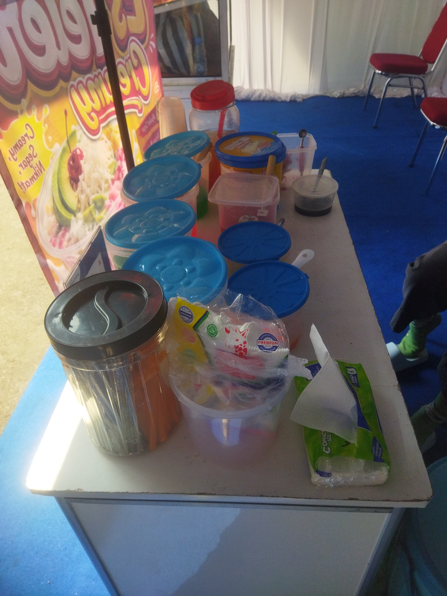 An outdoor food stall is set up in Giripurwo, Indonesia. A white table is covered with various items, including a tall clear plastic container filled with individually wrapped items, several blue-lidded containers, and a small white bowl with a dark substance and a spoon. To the left of the table is a large banner advertising a food item, possibly shaved ice or a similar dessert, with images of avocado and cherries. In the background, two red chairs with chrome legs are visible, set up on a blue carpet. A white curtain hangs in the background. The lighting suggests it is daytime, with bright sunlight casting reflections on the table surface.