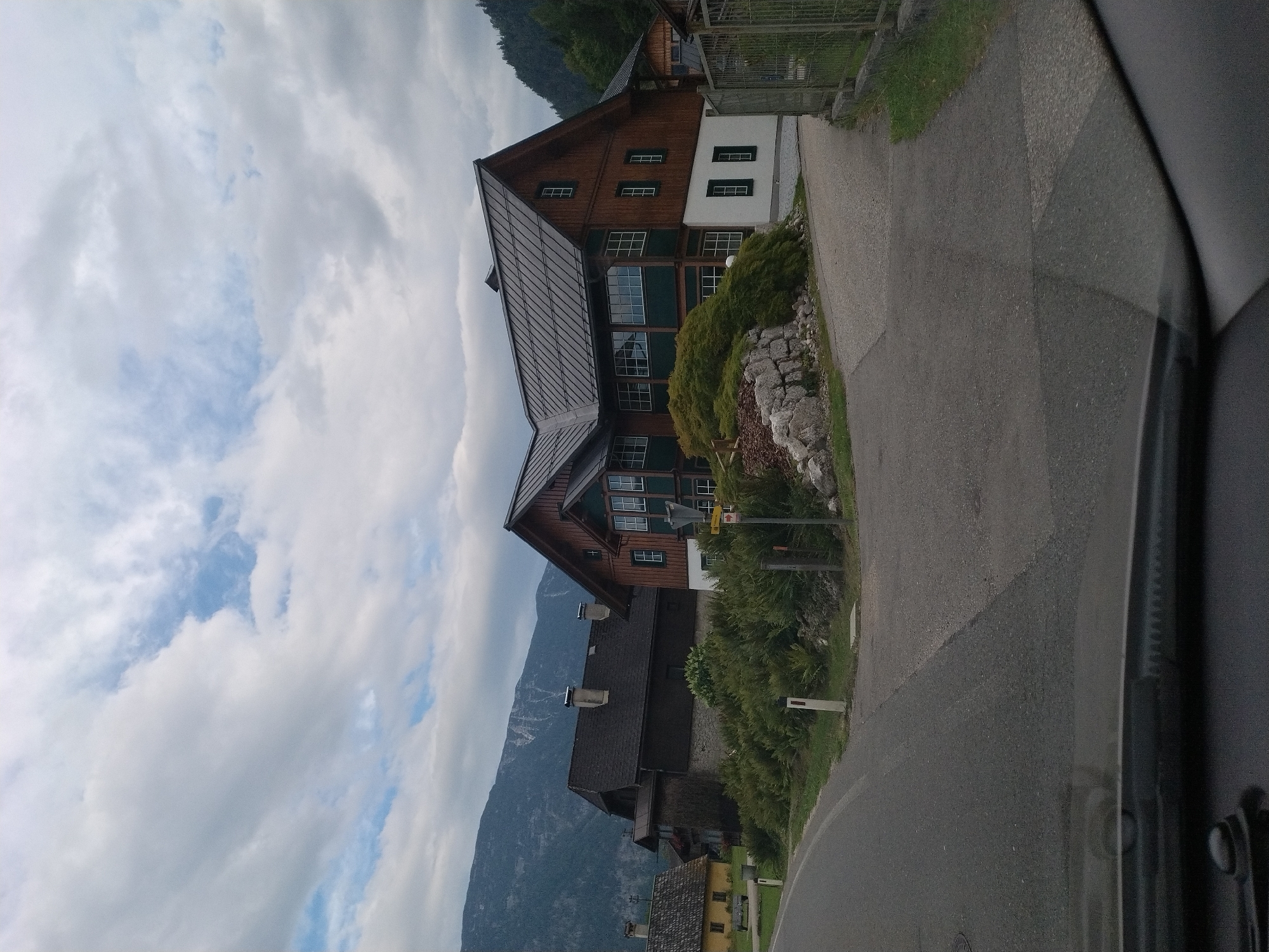 The image shows a rural landscape in St. Agatha, Austria, with a cloudy sky overhead and a mountainous backdrop. In the foreground, a paved road curves to the left. To the right of the road, a large, traditional Austrian house with a dark metal roof and wooden siding stands prominently. The house features multiple windows, some with dark green shutters. Adjacent to this house, a smaller, lighter-colored building is visible. To the left of the main road, another house with a yellow facade and a dark roof is situated. The area is surrounded by lush green vegetation and trees. A metal fence runs along the right side of the scene. The overall impression is one of a serene, picturesque village setting.