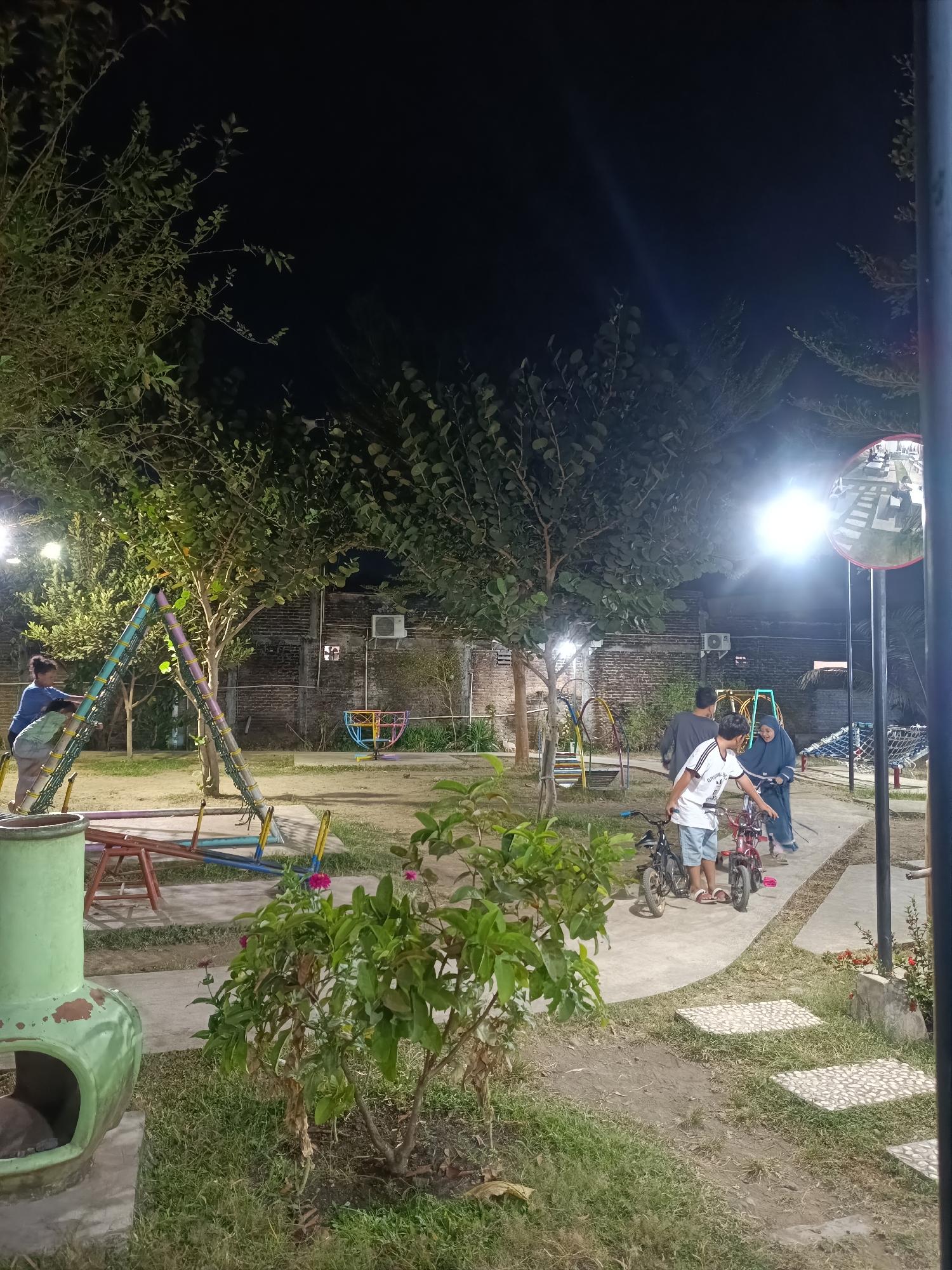 This image depicts an outdoor playground at night in Yogyakarta, Indonesia. In the foreground, a green, chimney-like object is situated next to a lush green bush with pink flowers. A paved pathway curves through the grass, leading to a seesaw-like playground structure. To the left, two young children are on a swing set. Further back, three individuals are standing near two bicycles. One person is wearing a white t-shirt and blue shorts, another a grey t-shirt, and the third a blue hijab. The background features a brick wall with an air conditioning unit and trees. A large, circular mirror on a pole is visible to the right, reflecting the illuminated playground. The scene is lit by artificial lights, creating a nighttime ambiance.
