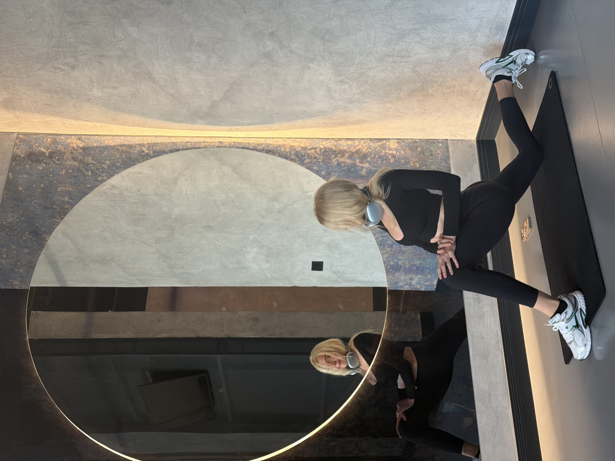 The scene depicts a modern fitness or studio interior in Belgrade, Serbia. A blonde woman, wearing a black long-sleeved crop top, black leggings, and white and green athletic shoes, is performing a low lunge stretch on a black yoga mat. Silver over-ear headphones rest around her neck. Her hands are clasped on her bent right knee, and her left leg is extended backward with the top of the foot flat on the mat. She is looking towards her reflection in a large, circular mirror that dominates the left side of the frame. The mirror features an internal, glowing light strip. The immediate background behind her consists of textured walls: a dark section with metallic flecks adjacent to the mirror, and a lighter grey, stucco-textured wall to the right. A dark baseboard runs along the bottom of the walls. The floor is dark grey. The mirror's reflection shows her face looking forward, along with reflections of a dark interior space containing what appears to be a mounted television or screen and dark vertical structural elements. A mobile phone with a colorful case is partially visible on the mat beside her right knee.