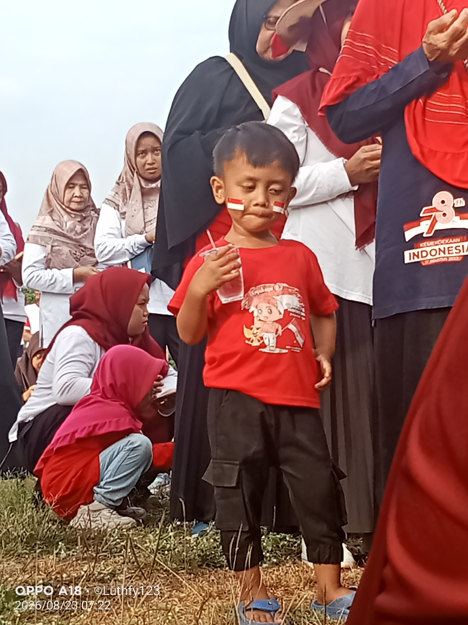 The image depicts a gathering of people, likely in an outdoor setting in Cawas, Indonesia, given the timestamp and watermarks. The central subject is a young boy wearing a red t-shirt with a cartoon character and Indonesian flag colors on his cheeks. He is holding a cup. He is surrounded by several individuals, mostly women, wearing headscarves and traditional attire in various colors like red, beige, and white. The clothing suggests a festive or commemorative event, possibly related to Indonesian Independence Day, as indicated by the "78 INDONESIA" graphic on a dark blue shirt visible in the background. The ground is covered with dry grass. The overall scene conveys a sense of community and celebration.
