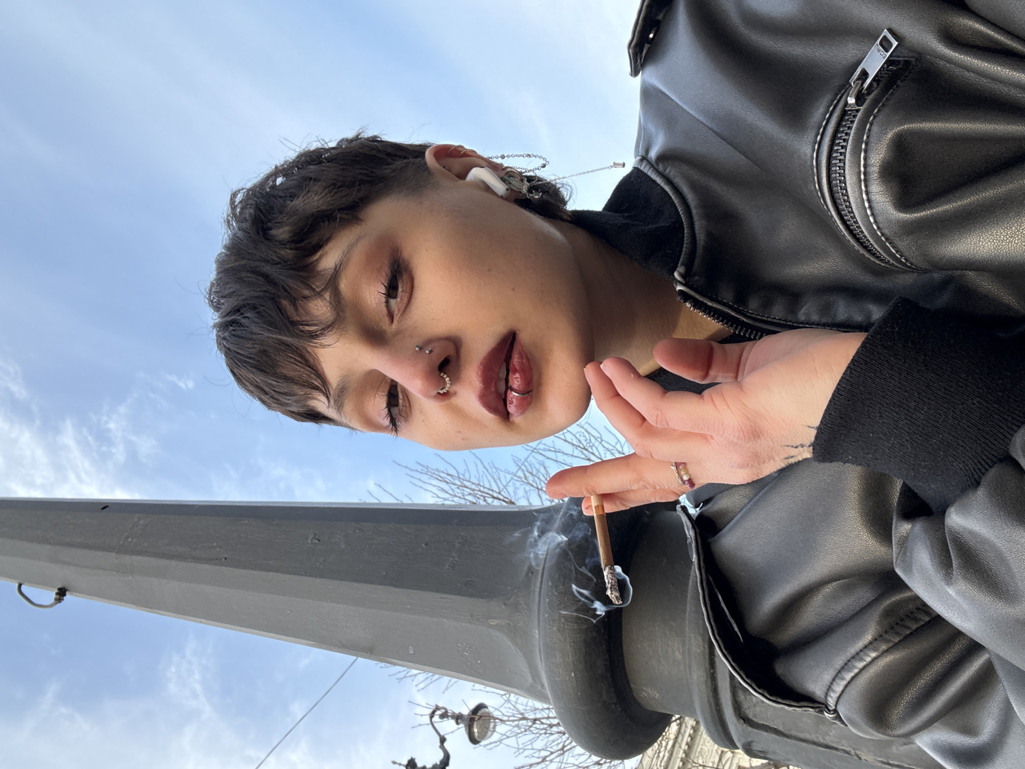 A low-angle shot depicts a person with short, dark hair and multiple piercings (nose, lip) holding a lit cigarette in their right hand. The person wears an AirPod in their right ear, adorned with a chain. They are wearing a black leather jacket over a black long-sleeved shirt. The cigarette is emitting smoke, and is propped on a gray, rounded metal object (potentially a lamp post base). A building and tree branches are partially visible in the background, against a blue sky with scattered clouds. Given the architectural details visible, the location is plausibly in Saint Petersburg, Russia.