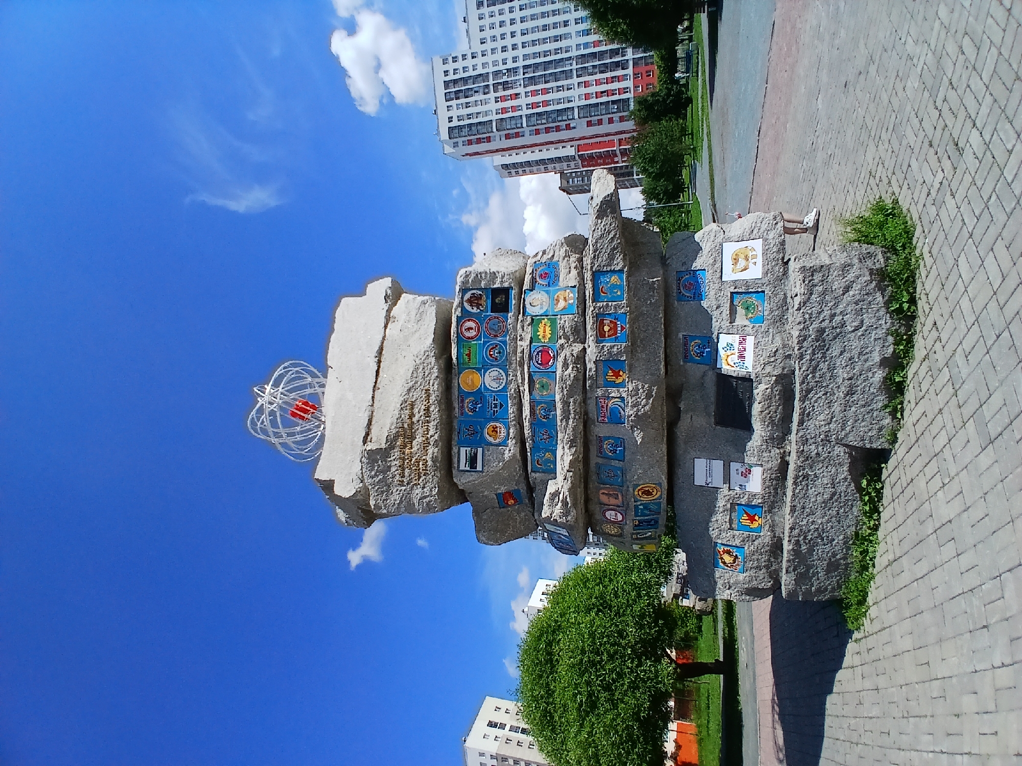 A stacked stone monument adorned with colorful tiles and a wire sculpture topped with red accents stands in an urban park on a sunny day. The monument is surrounded by a paved walkway and green grass. In the background, modern apartment buildings rise against a bright blue sky with scattered clouds. A large, green tree is visible to the left of the monument, and a portion of a person's legs and feet in white sneakers is visible to the right, suggesting they are standing near the monument. The monument appears to be a public art installation, likely located in the city of Yekaterinburg, Russia, given the stylistic elements of the architecture and the monument itself.