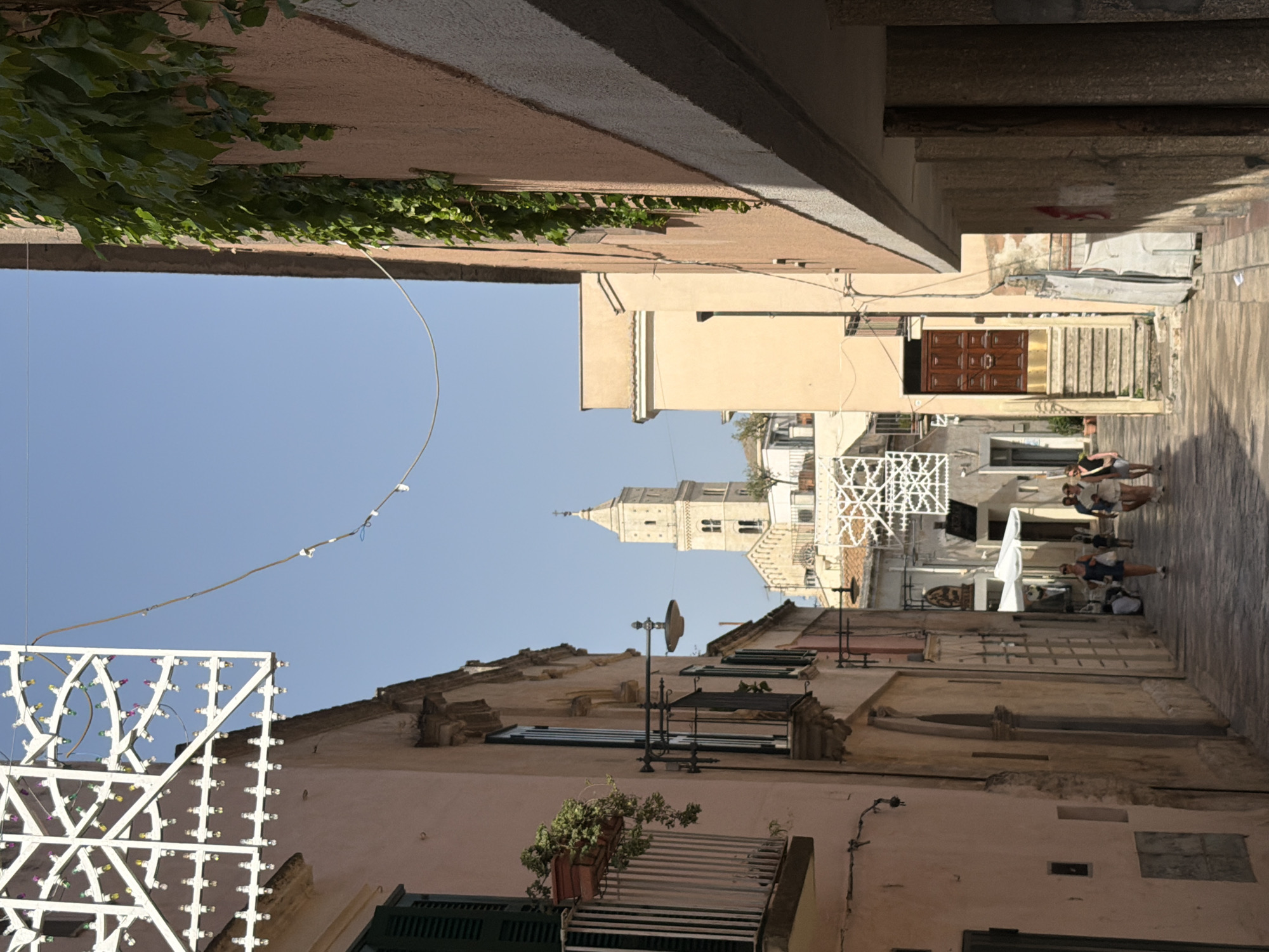 The image captures a view down a narrow, cobblestone street in Matera, Italy, under a clear blue sky. On the left, a pale pink building features a balcony with a potted plant and a closed dark green shuttered window. Further down, another building with similar architectural style has a balcony and a street lamp. On the right, a peach-colored building with ivy growing on its facade has a section of its exterior shaded. In the distance, a tall, pale stone bell tower of a church dominates the skyline, with a white, decorative structure and some buildings with terracotta roofs visible below it. Several people are walking down the street; a woman in a blue dress and a child are in the middle ground, followed by a group of three people further down. The street is lined with aged buildings, some appearing to be made of stone, with elements of wear and tear visible. The overall scene conveys a sense of an old, Mediterranean town.