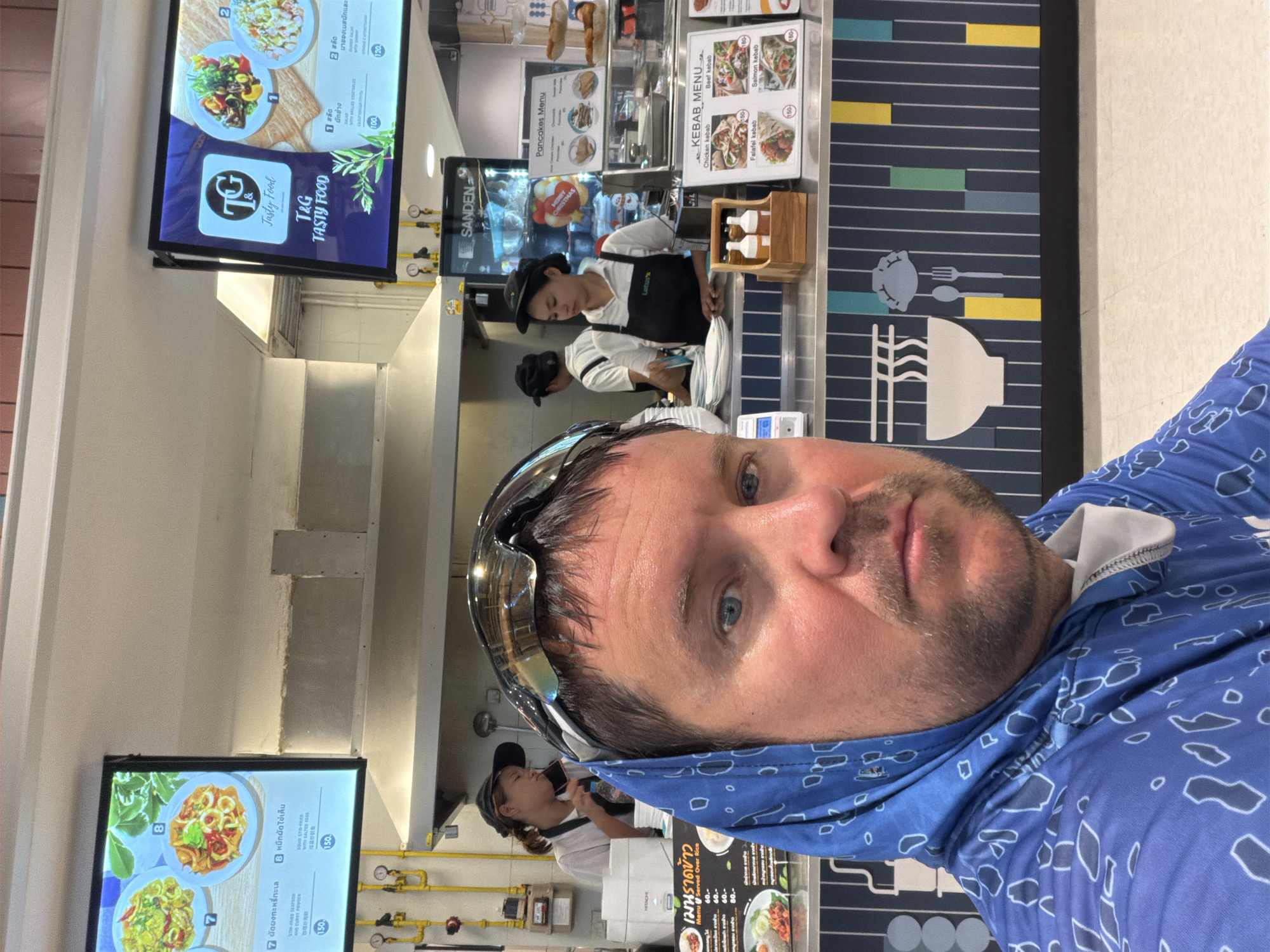 A man wearing a blue hooded sweatshirt and sunglasses is taking a selfie in front of a food stall. The food stall has menus displaying various dishes, including "Stir-fried seafood and curry powder" and "Squid stir-fried with chili paste." Behind the counter, several employees are visible, including two women in black aprons and a man in a white shirt and black cap. The stall features a "Pancakes Menu" and a "Kebab Menu" with pictures of chicken, beef, falafel, and salmon kebabs. The background shows a food court environment with tiled walls and lighting fixtures. The image appears to be taken in Ban Bang Rak, Thailand, as suggested by the presence of Thai text on the menus.