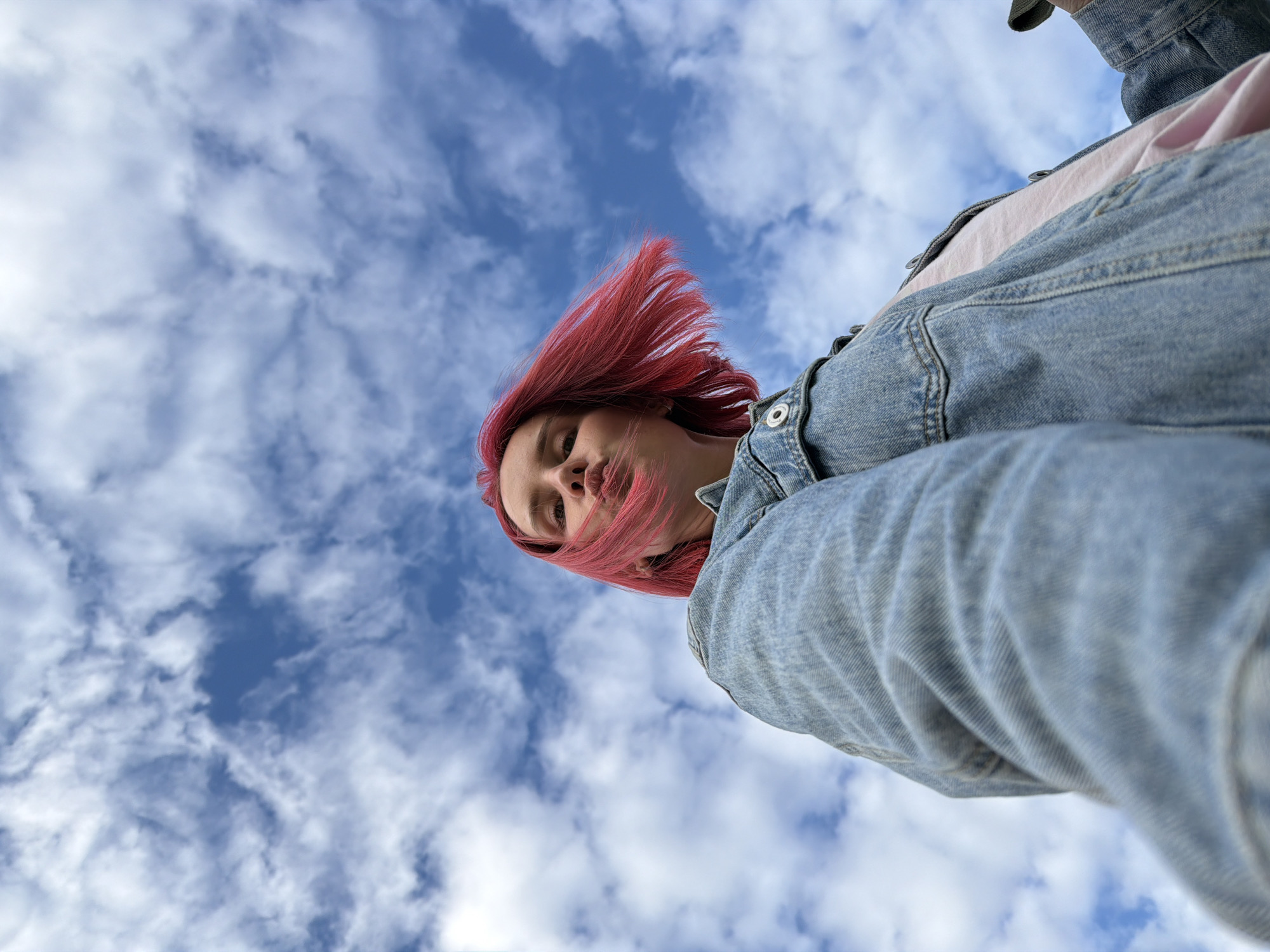 A low-angle, close-up, Dutch angle, medium shot captures a person with bright pink hair, wearing a denim jacket and a pink t-shirt, looking up towards the sky. The person's face is positioned in the upper center of the frame, with their lips puckered. The background consists of a bright blue sky filled with scattered white clouds. The image appears to be a selfie taken outdoors. There is no indication of the specific location within the city of Chaikovsky, Russia.