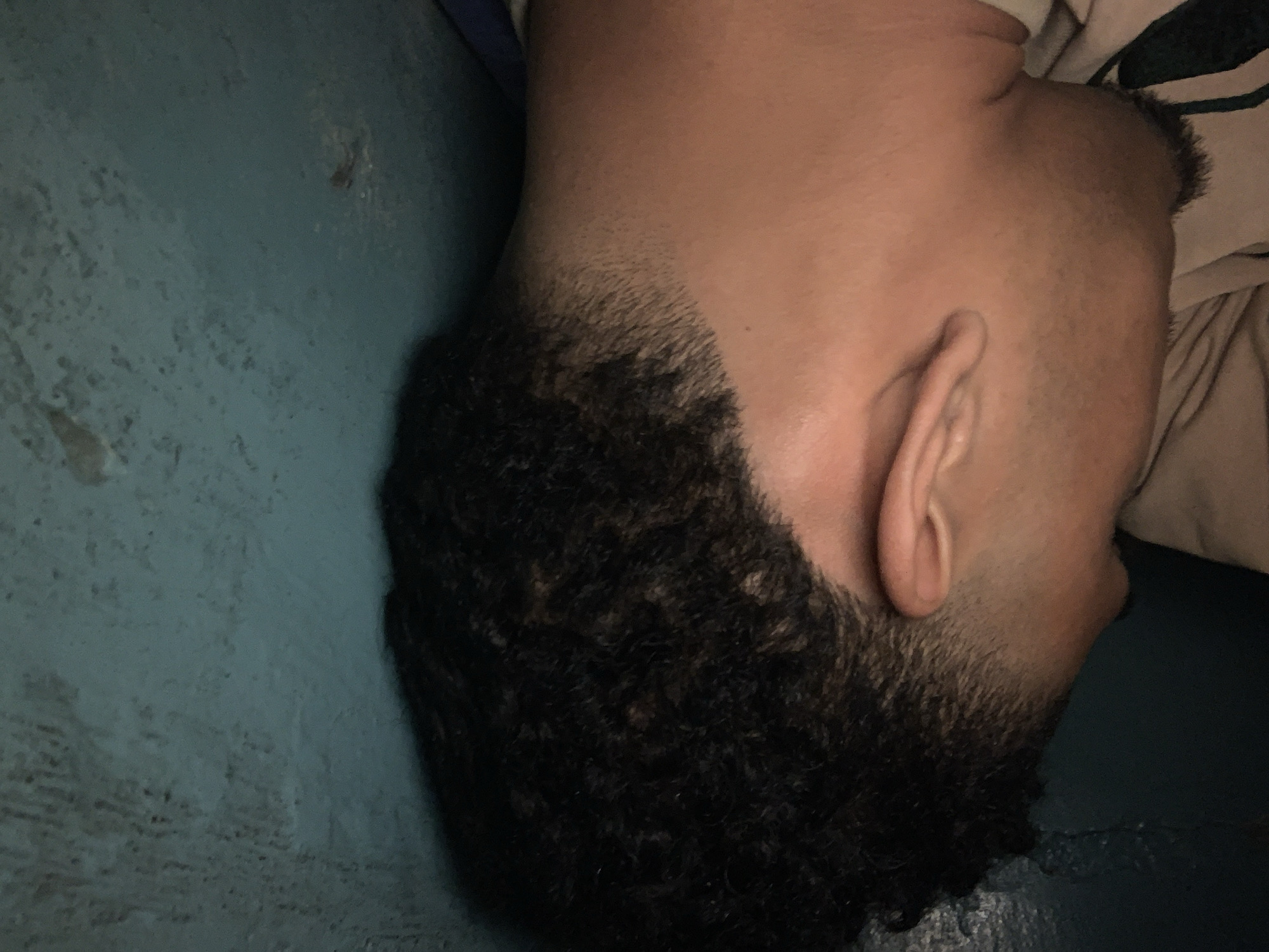The image captures a close-up of a person's head, oriented sideways, seemingly resting against a surface. The subject is an individual, likely male, with short, dark, curly hair styled with a distinct fade on the sides. The skin tone is medium. Their ear is prominently visible on the right side of the frame, and stubble can be seen along the jawline and sideburn area, indicating recently trimmed facial hair or a very close shave. A small mole is visible on the side of the neck/jawline.
The person appears to be in a state of rest, possibly lying down or with their head tilted against a surface. A light-colored fabric, which could be part of a shirt or bedding, is visible near the person's neck and shoulder.
The background and foreground consist primarily of a muted blue-green surface, which exhibits a slightly textured appearance, suggesting it could be a painted wall, a floor, or a piece of furniture. The lighting is dim and soft, creating a subdued atmosphere, and does not allow for a clear determination of the time of day, but suggests an indoor setting in Santa Ana, Venezuela. There is no visible text in the image, and no specific emotions can be discerned from the person's visible features.