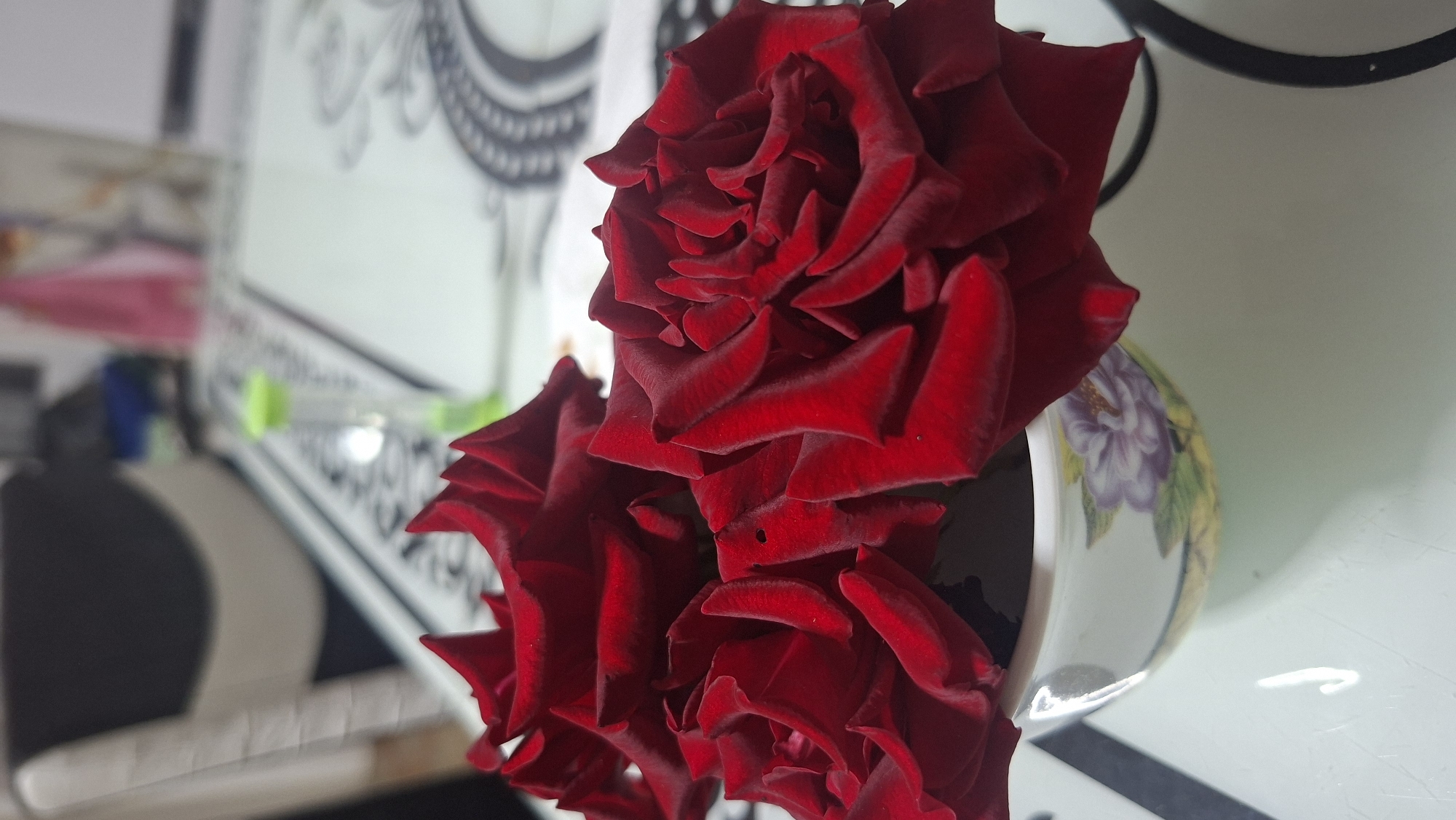 A close-up shot focuses on a bouquet of three deep red roses with velvety petals, partially obscuring a small, decorative ceramic pot with a floral design. The pot rests on a patterned tablecloth, which covers a table. In the blurred background, a chair with a dark backrest and a lighter seat is visible, along with hints of other objects and possibly a window. The scene is indoors, likely a home setting in Voinova, Moldova.