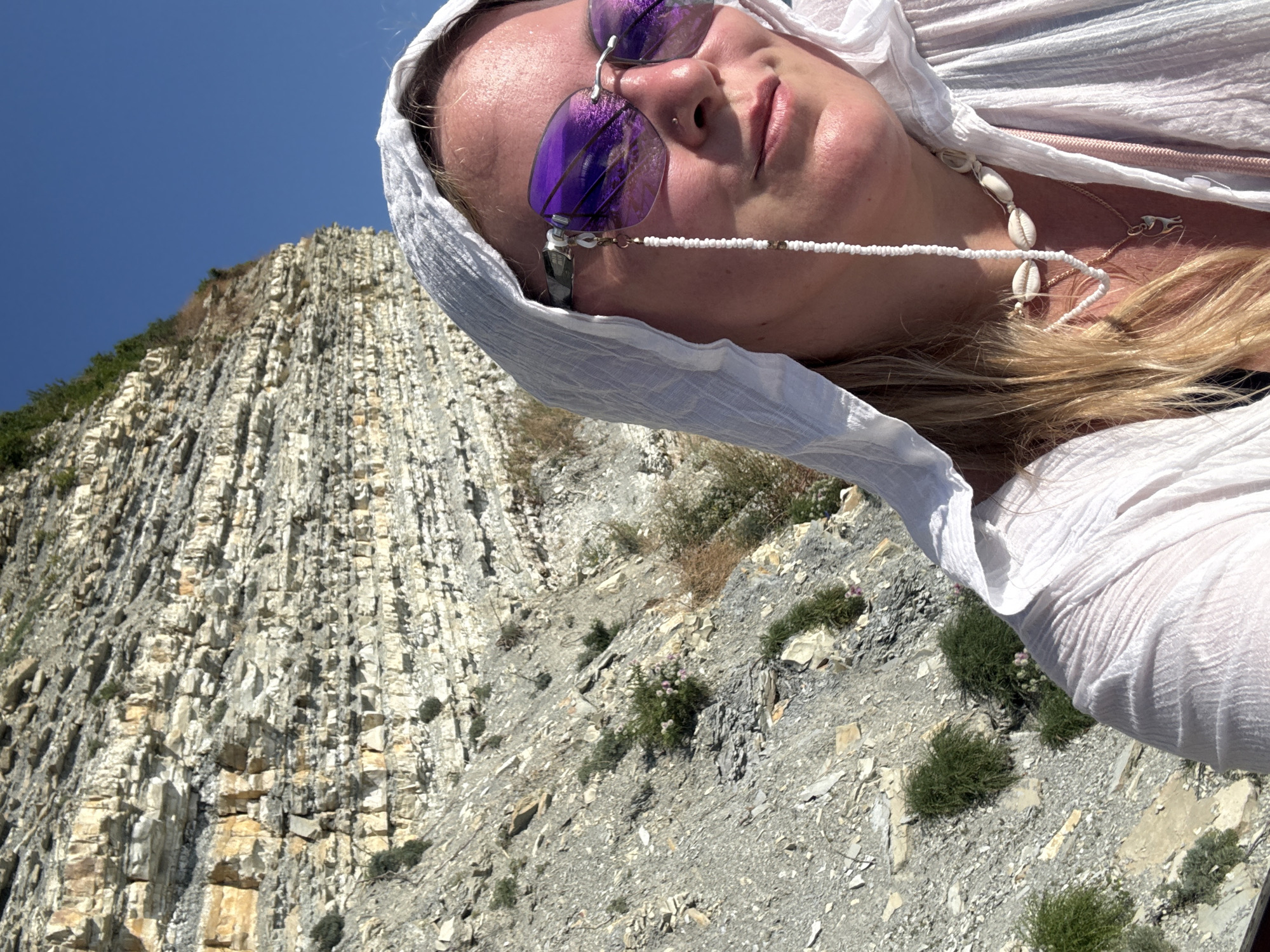 A medium shot captures a woman in a white hooded garment and sunglasses, posing for a selfie with a rocky cliff face and a clear blue sky in the background. She wears a white beaded eyeglass chain adorned with seashells and a silver cat pendant. The location is the city of Kabardinka, Russia, specifically the rocky shores of the Black Sea. The cliff face exhibits layered sedimentary rock formations, with patches of sparse vegetation. The lighting suggests it is daytime, with the sun casting reflections on the woman's sunglasses.