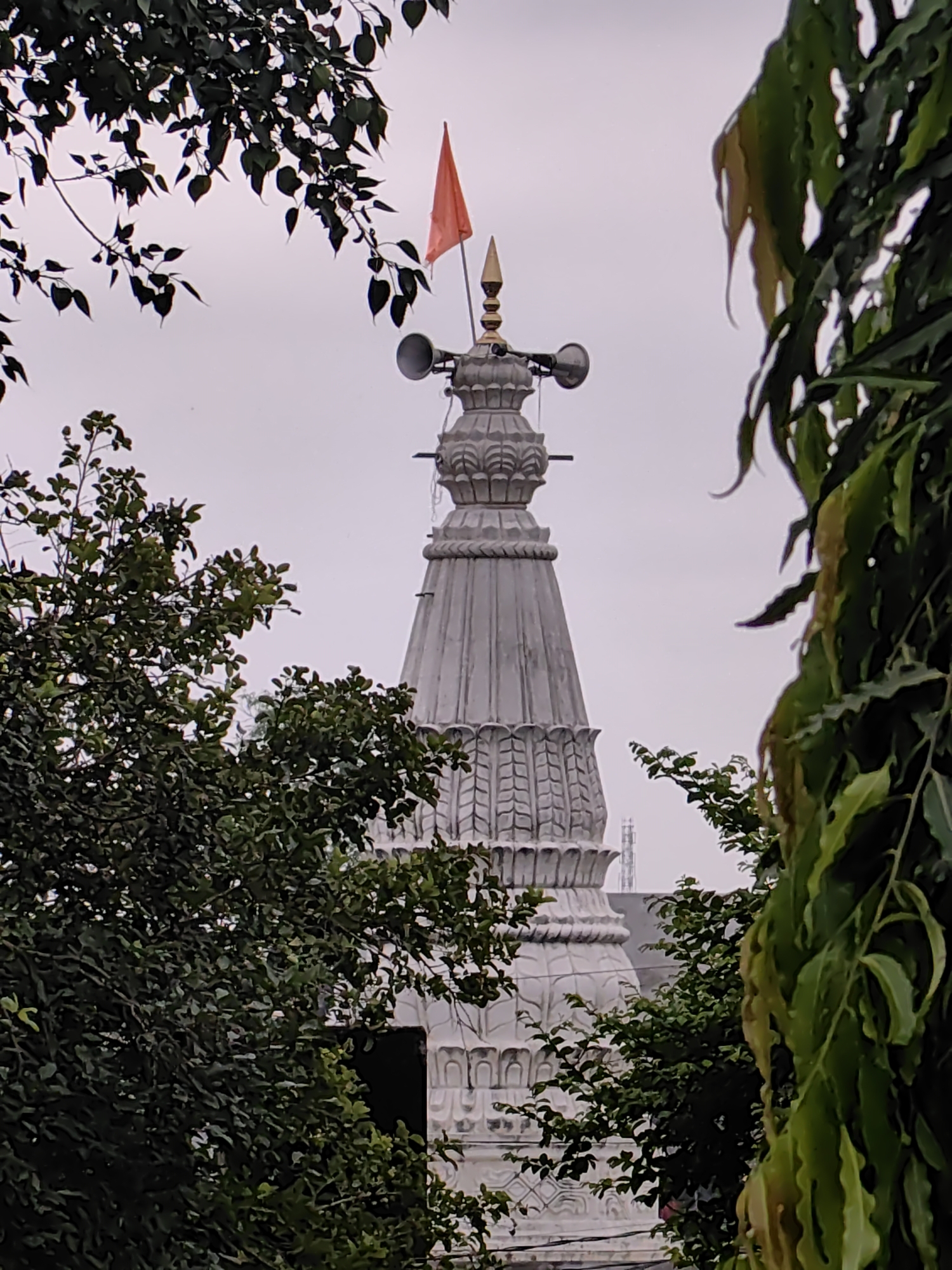 The image shows a white spire of a temple with a red flag on top. The spire is ornately decorated with carvings and has two loudspeakers mounted on it. The temple is partially obscured by lush green trees with dark leaves. The background is a hazy, light gray sky. In the distance, a cell tower can be seen behind the temple. The image appears to be taken in Mhaiswadi, India. No people are visible in the image.