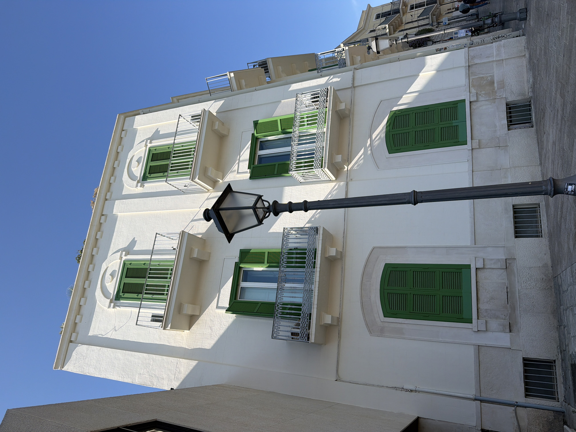 This image depicts a white building with green shutters on its windows and balconies, illuminated by sunlight under a clear blue sky. The building is located in the city of Bari, Italy, specifically in the historic center, near the waterfront. A street lamp stands in the foreground, casting a shadow on the building and the cobblestone street. The architecture is typical of the region, with balconies adorned with intricate metal railings. The scene is tranquil, with no people actively engaged in any discernible activity, though two figures can be seen in the distance on the right side of the image, suggesting the area is frequented by pedestrians. The overall impression is one of a sunny, picturesque European streetscape.