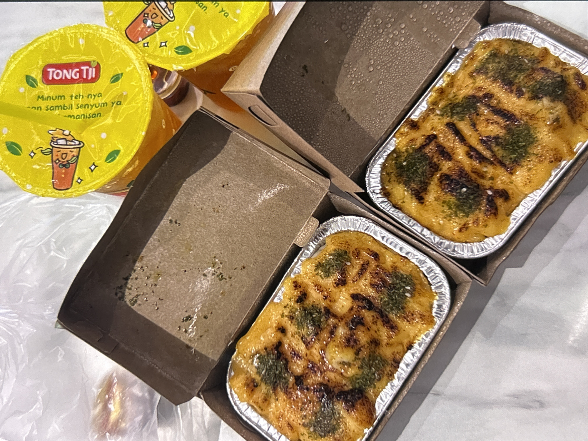 Two foil containers filled with baked food, likely some type of casserole or gratin, are presented in open cardboard boxes. The food is topped with a golden-brown sauce and sprinkled with green herbs. Two yellow plastic cups with red and white branding that says "Tong Tji" are visible in the background. The scene appears to be a casual meal, possibly takeout or delivery, with a white marble-patterned surface underneath. The lighting suggests an indoor setting. The image is taken from a top-down perspective. The presence of "Tong Tji" branding strongly suggests the beverages are from that popular Indonesian tea brand, and Depok, Indonesia is known to have numerous Tong Tji outlets.