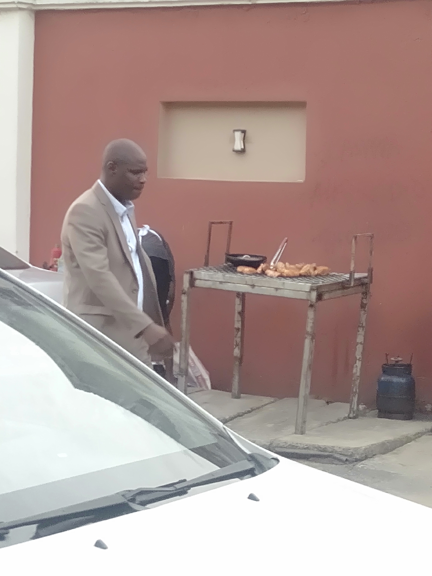 The image shows a man in a tan suit walking past a white car and a metal grill with food on it. The grill is located against a building with a red wall and a recessed, beige rectangular area with a white fixture inside. A propane tank is visible next to the grill. The location is identified as Unknown, Unknown.