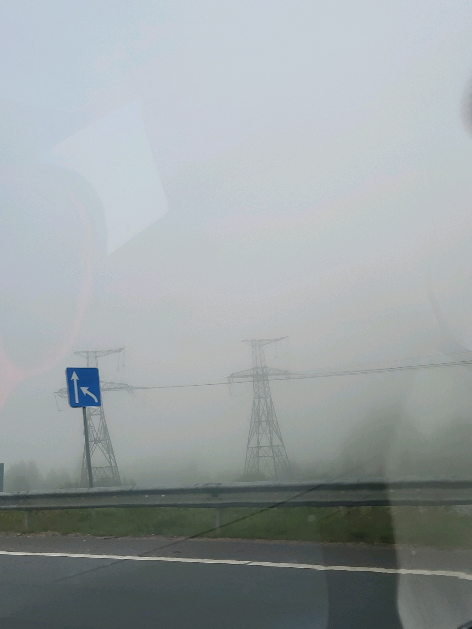 The image depicts a foggy day in Brankas, Latvia, with visibility significantly reduced. In the foreground, a road can be seen, with a metallic guardrail separating it from a grassy area. A blue traffic sign with white arrows indicates that traffic can proceed straight ahead or turn left. Beyond the guardrail, two large electricity pylons are visible, their structures partially obscured by the dense fog. The pylons are spaced apart, suggesting they are part of a power line transmission system. The overall atmosphere is muted, with the fog creating a sense of isolation and obscuring distant details. There are no people or vehicles clearly visible in the scene.