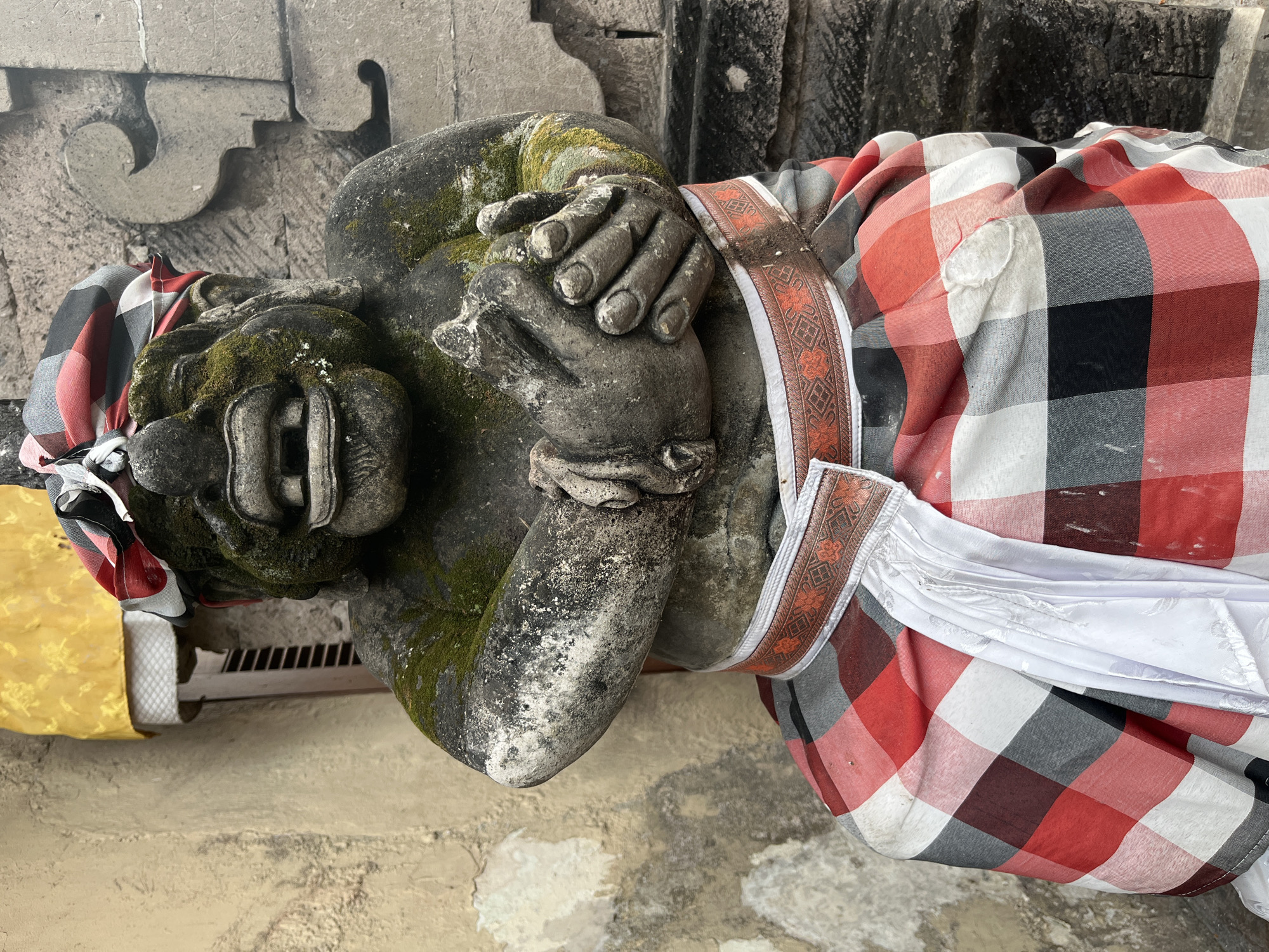 A stone statue of a grotesque figure, possibly a guardian, is seated in front of a textured, tan-colored wall. The statue is adorned with moss, giving it a weathered appearance. It wears a headdress with red, black, and white checkered patterns, and a sarong of the same pattern around its waist. The sarong is accented with an orange and white patterned sash. The figure's hands are clasped together in front of its chest, and its mouth is open in a wide, toothy grimace. To the left of the statue, a yellow fabric is draped over what appears to be a wooden structure with louvred openings. The background consists of carved stone elements and a darker, rougher stone wall. The scene is outdoors, with natural light illuminating the statue and its surroundings. The location is identified as Kuta Selatan, Indonesia.