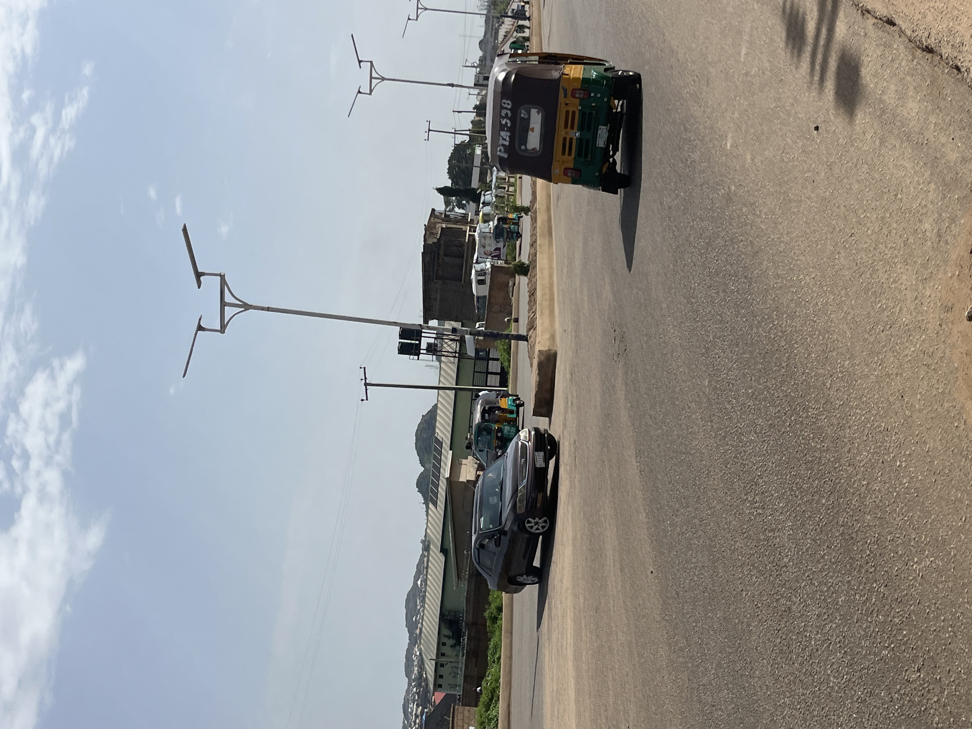 In Bukuru, Nigeria, a dark-colored sedan drives on a paved road. To the right of the sedan, a green and yellow auto-rickshaw with license plate "PTA 558" is visible. In the background, buildings and light poles line the road. The sky is partly cloudy.