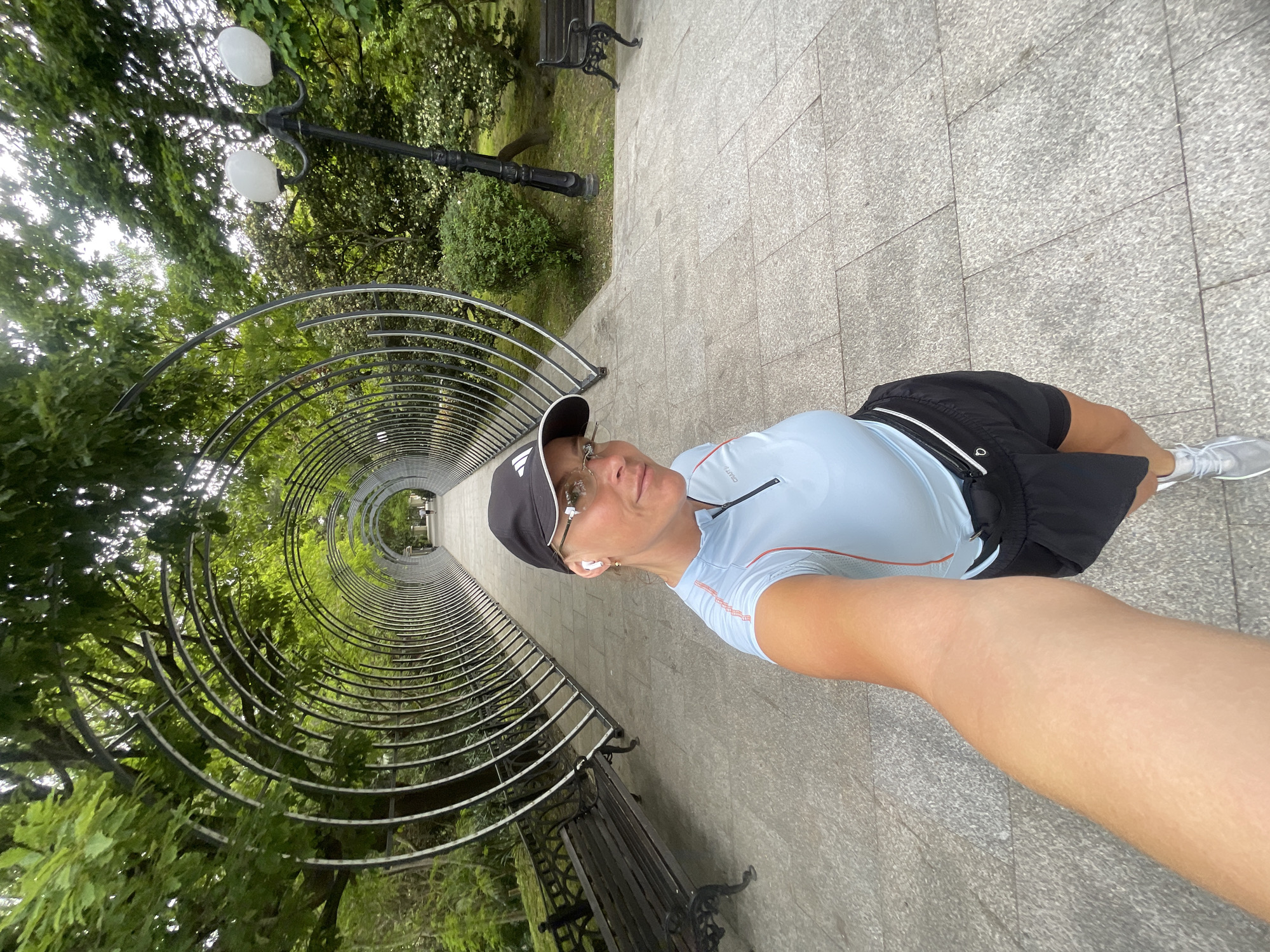 A female is taking a selfie in Sochi, Russia. She is in a park, on a paved path, with a curved metal structure forming a tunnel overhead. The path has benches on either side. The trees are lush with green leaves. There is a lamp post visible in the background. The woman is wearing a baseball cap, sunglasses, athletic clothing, and earbuds. She is holding the camera out with her arm.