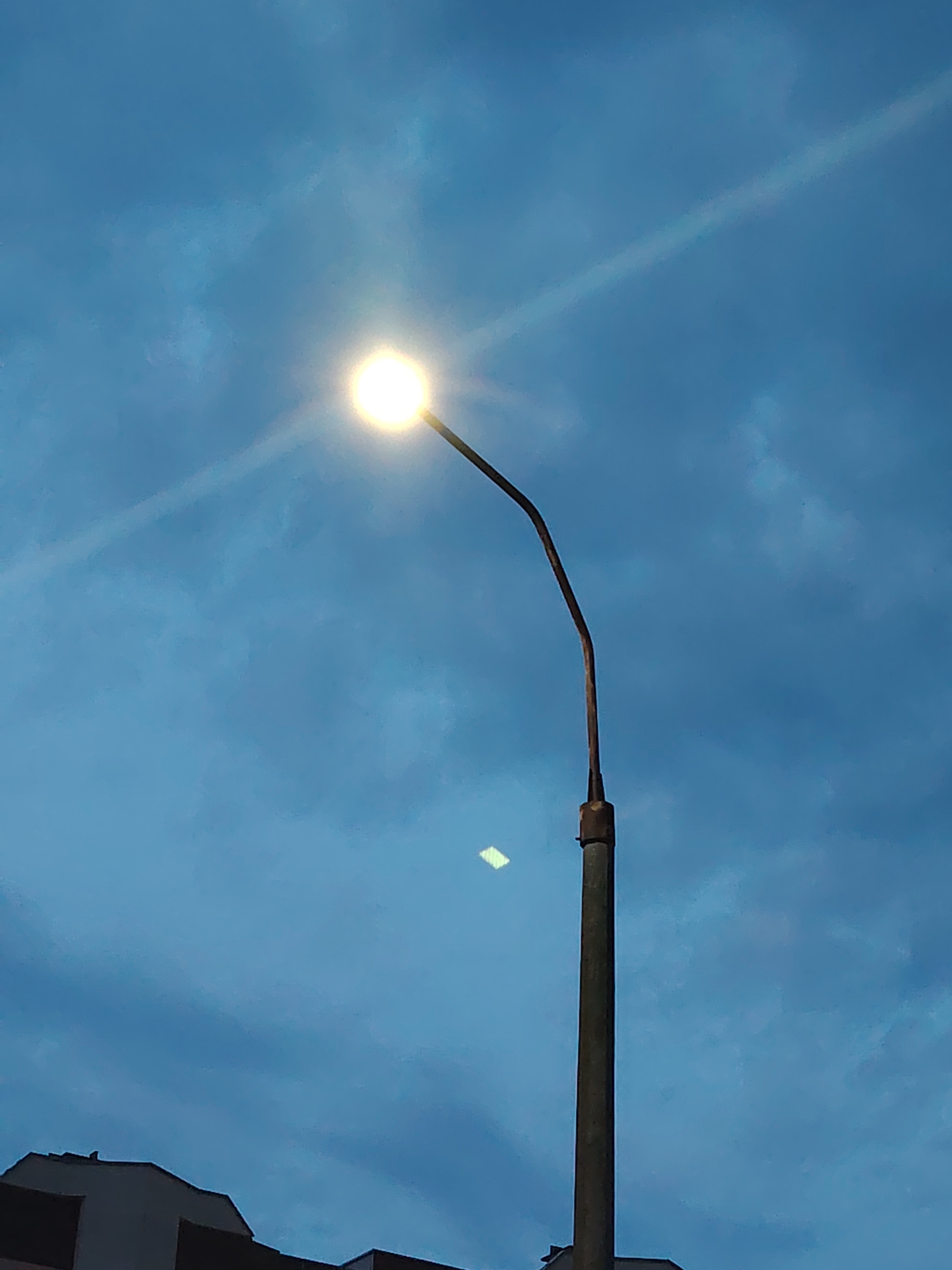An outdoor, low-angle shot captures a streetlamp against a predominantly blue sky with scattered clouds. The streetlamp's light is a bright, luminous circle, with a visible beam or lens flare extending diagonally across the frame from the upper left to the lower right. The lamp post is a weathered, vertical pole with a curved arm supporting the light. In the lower left corner, the dark silhouette of a building or roofline is visible, suggesting an urban or suburban setting. The sky exhibits a twilight or dusk hue, with the clouds appearing somewhat textured and muted. A faint, rectangular, bright reflection is present on the sky, to the left of the lamp post. The location is specified as Baravlyanski Sel'ski Savet, Belarus.