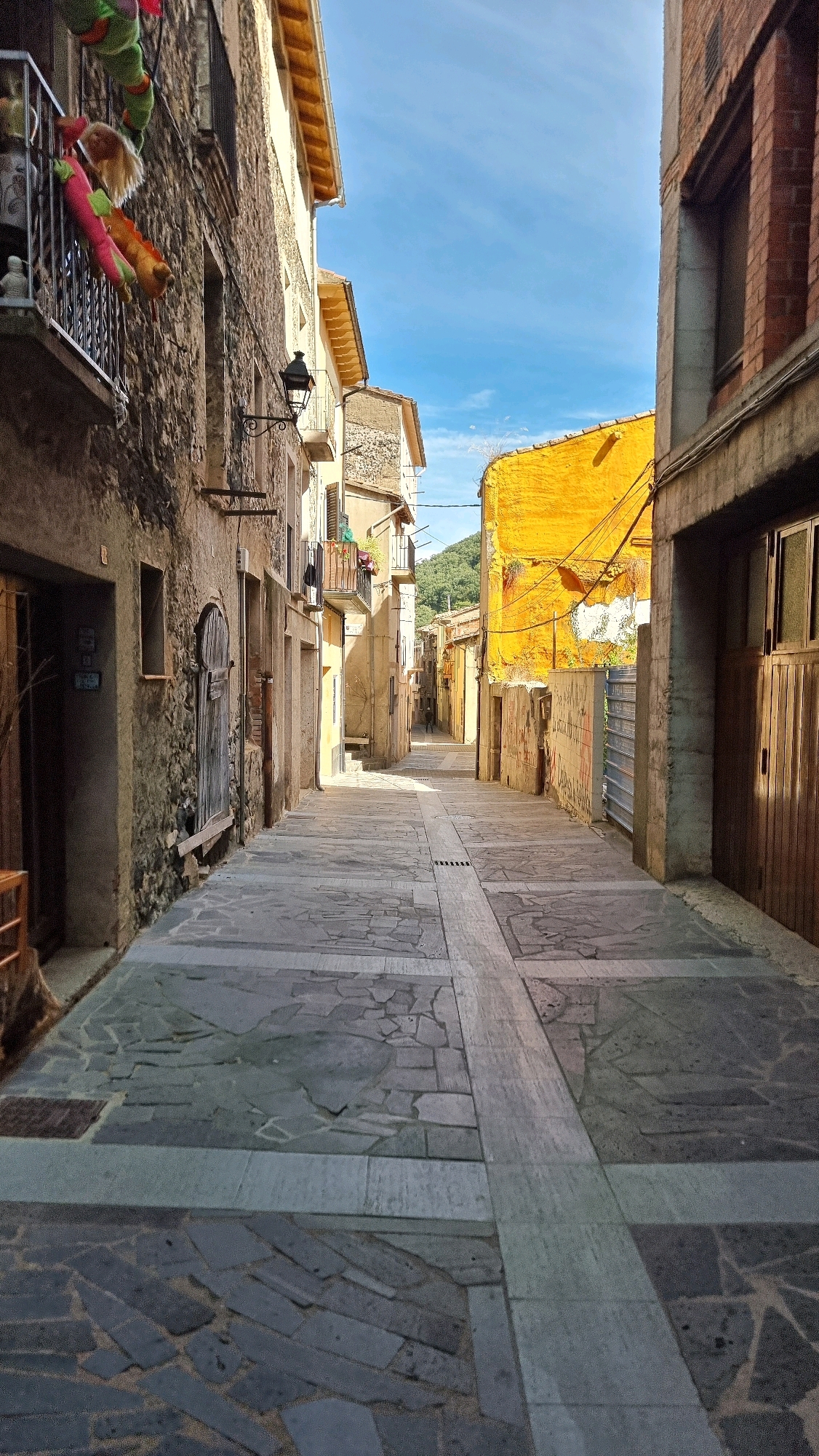 The image depicts a narrow, cobblestone street in the town of Castellfollit De La Roca, Spain. The street is lined with stone and yellow stucco buildings, some with balconies adorned with colorful stuffed toys. The sky is a clear blue with some scattered clouds. Sunlight casts shadows on the street, indicating it is daytime. A single person is visible walking in the distance, down the street. The architecture suggests a historic European town.