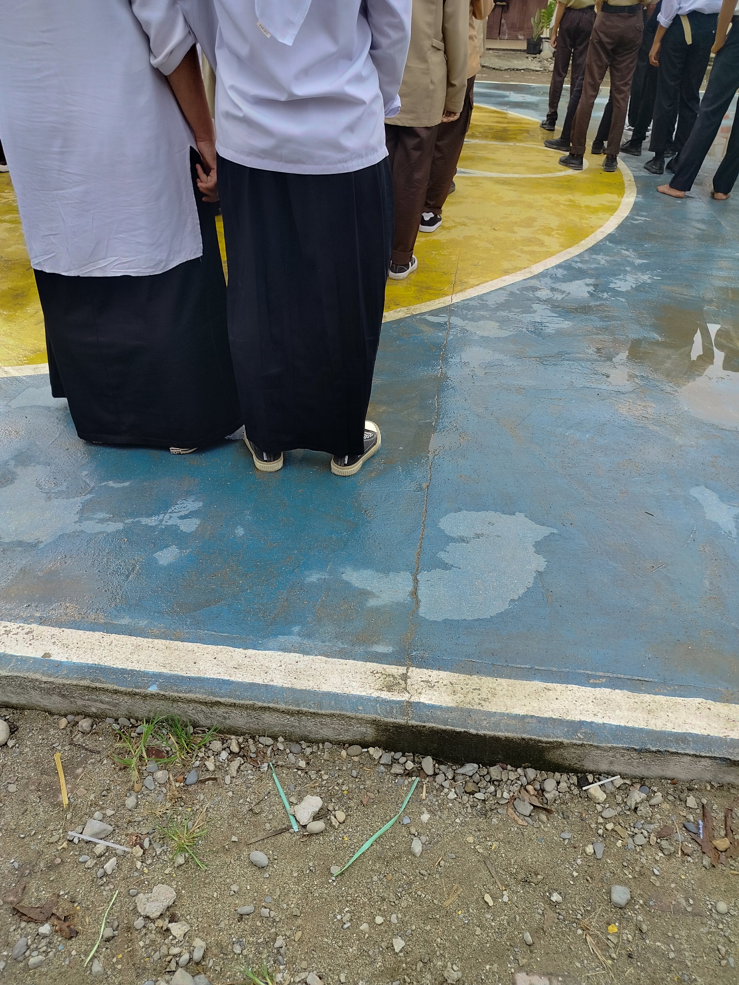 In Lawebekung, Indonesia, a group of people, likely students given their uniforms, are standing on a wet, blue and yellow basketball court. Two individuals in the foreground are wearing white shirts and long black skirts. One of them is wearing black sneakers with white soles and laces, while the other is also wearing dark-colored sneakers. Behind them, a line of people in tan jackets and brown pants, along with others in dark uniforms, stand facing away from the camera. The court surface shows puddles of water and reflections, indicating recent rain. The ground surrounding the court is a mix of sand, small stones, and sparse vegetation. The scene appears to be an outdoor gathering or activity on the court.