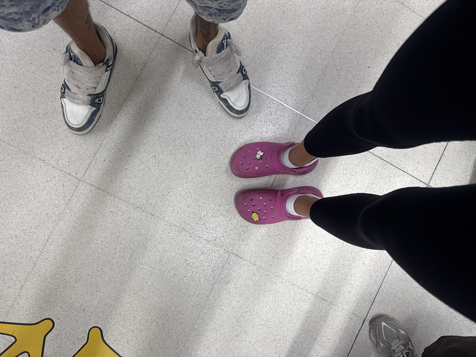The image captures an overhead view of several people's feet and lower legs on a light grey, speckled tiled floor, likely in an indoor public setting such as a mall or transportation hub in Tambon Mai Khao, Thailand.
In the upper left, a person is seen wearing what appears to be faded blue shorts or jeans, paired with blue and white sneakers featuring distinctive thick, fluffy white laces. A tattoo is partially visible on their lower leg. To the right, another individual wears bright pink Crocs with white socks and black ribbed leggings. One of the pink Crocs is adorned with a yellow Jibbitz charm, which appears to be a cartoon character. These Crocs show some signs of wear and minor dirt. In the bottom right corner, the feet of a third person are partially visible, wearing grey athletic shoes and dark-colored pants.
The floor is consistent throughout, characterized by its light grey color and speckled texture, with visible tile seams and a thin, metallic-looking expansion joint running horizontally across the mid-ground. In the bottom left, a portion of a yellow outline or graphic is visible on the floor, suggesting a designated area or decorative marking. The people appear to be standing or casually moving through the space. There is no discernible text visible within the frame. The lighting indicates an indoor environment.