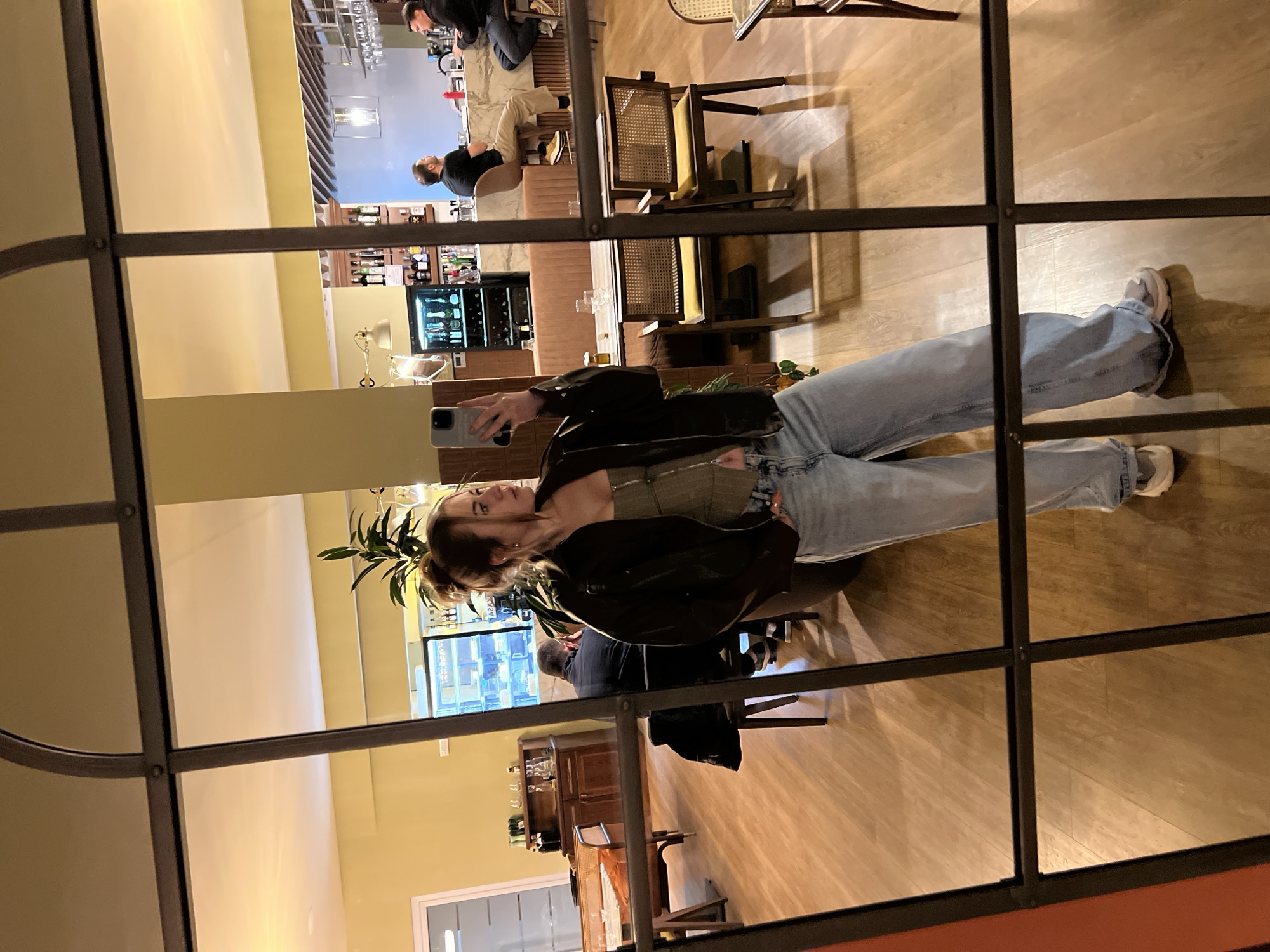An interior shot depicts a young adult female taking a mirror selfie within a restaurant or bar in Boreti, Montenegro. The female subject, positioned centrally, wears a dark jacket, an olive-toned top, light-wash wide-leg jeans, and light sneakers. Her light brown hair is styled in a bun, and she holds a light-colored smartphone with her right hand at chest level, capturing her reflection. Her left hand is partially in her left front pocket.
The mirror's dark, arched frame and grid-like dividers are visible in the foreground. The reflection reveals a dining establishment with warm ambient lighting. The walls are predominantly light yellow, transitioning to a light blue/grey area behind the bar. The flooring is light brown wood-patterned laminate.
The reflected interior features various dark wood tables, some set with glasses. Cane-back chairs with yellow upholstered seats are arranged around the tables. A long, light brown upholstered booth runs along a wall. A marble or light-colored stone bar counter occupies the right side of the reflection, with two adult males seated on dark brown bar stools; one wears a black shirt and beige pants, the other a black shirt and dark pants. Behind the bar, shelves display bottles, and a dark refrigerator/wine cooler is visible. A red squeeze bottle rests on the bar counter.
On the left side of the reflection, another adult male in a dark jacket is seated at a table, facing away. A dark jacket is draped over an adjacent chair. A dark wooden credenza with bottles and glasses is also present on the left. A large green potted plant stands near the bar area.