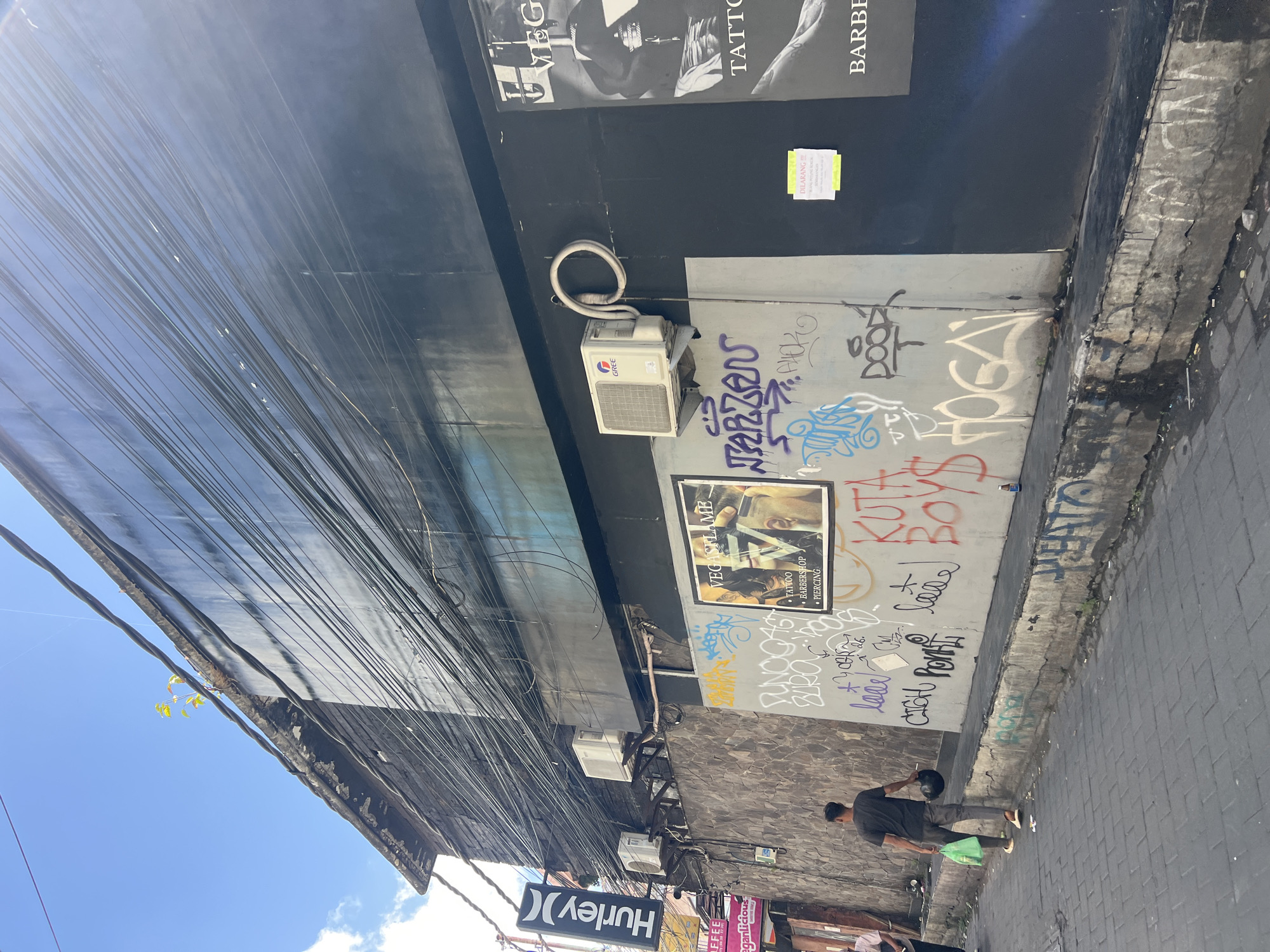 This image captures a street scene in Kuta, Indonesia, characterized by a predominantly dark building facade covered in a dense network of black electrical wires. The building appears to be a commercial establishment, with signage for "Hurley" visible on the left. Below the facade, a rough stone wall is plastered and adorned with various graffiti tags and a poster. The poster advertises "VEGAS FLAME" and lists services such as Tattoo, Barbershop, and Piercing.
In the foreground, a man dressed in a dark t-shirt and grey pants walks away from the camera, carrying a green plastic bag and a black helmet. He is walking on a paved sidewalk. The street is narrow, with a concrete curb running along the wall, also showing signs of wear and graffiti. There are a couple of air conditioning units mounted on the building above the stone wall. The sky is blue with a few scattered clouds. The overall impression is one of an urban environment with signs of commercial activity and street art.