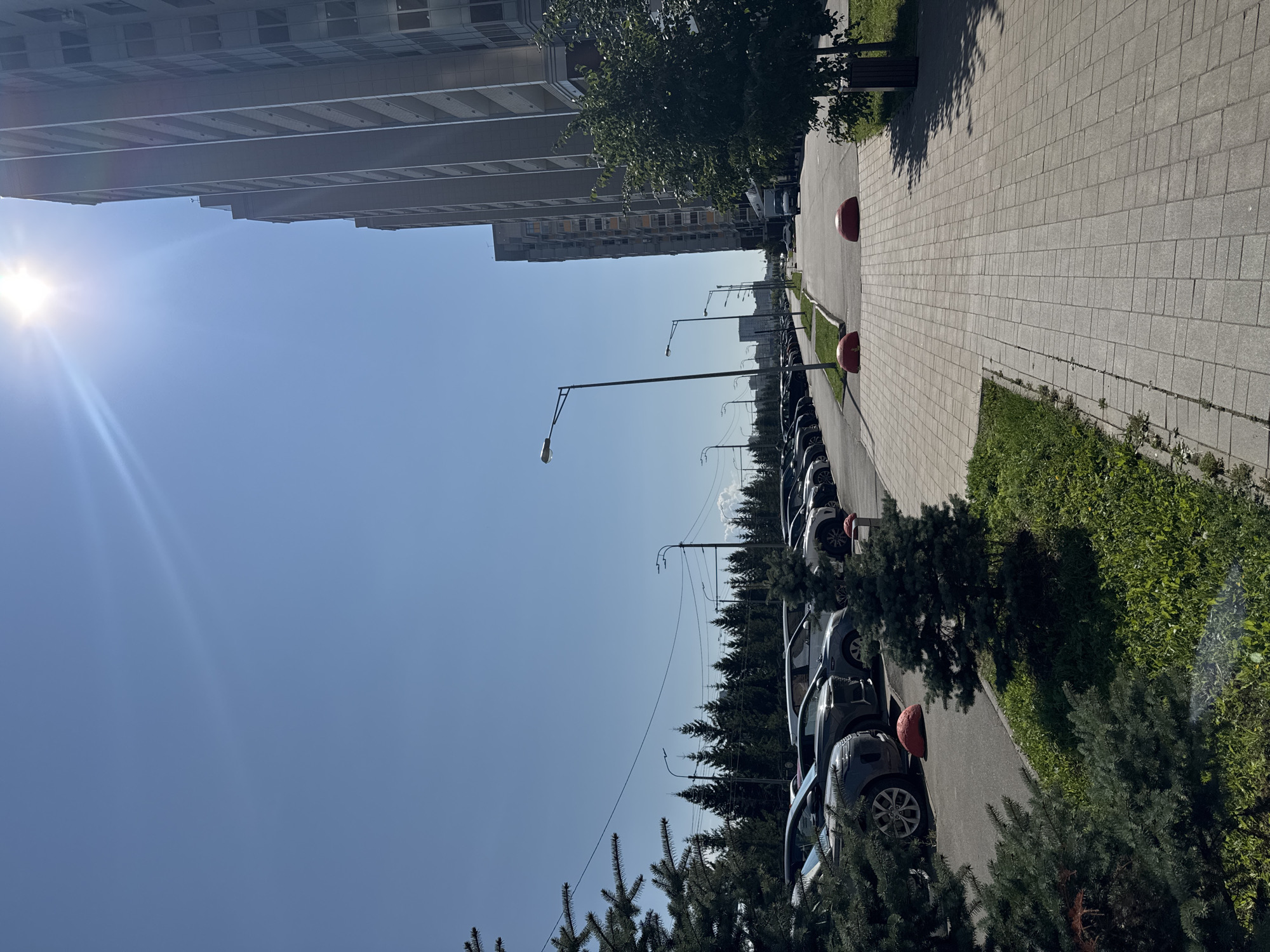The image captures a street scene in Kazan, Russia, on a clear, sunny day. On the right, a tall, modern residential building with multiple balconies dominates the skyline, its facade a light gray. The sun, a bright white orb, shines intensely in the upper right corner, casting lens flare across the image. In front of the building, a paved sidewalk is lined with green grass and a few trees. Two large, red, dome-shaped bollards are placed on the sidewalk, seemingly to prevent vehicles from entering the pedestrian area.
To the left of the sidewalk, a street runs parallel, lined with parked cars. The cars are of various models and colors, mostly dark, suggesting a parking lot. Beyond the cars, a dense line of coniferous trees creates a green backdrop. Further in the distance, more trees and buildings are visible under the bright blue sky. Streetlights are positioned along the street, one of them prominently in the mid-ground. The overall scene depicts a well-maintained urban environment, likely a residential area, with ample parking and green spaces.