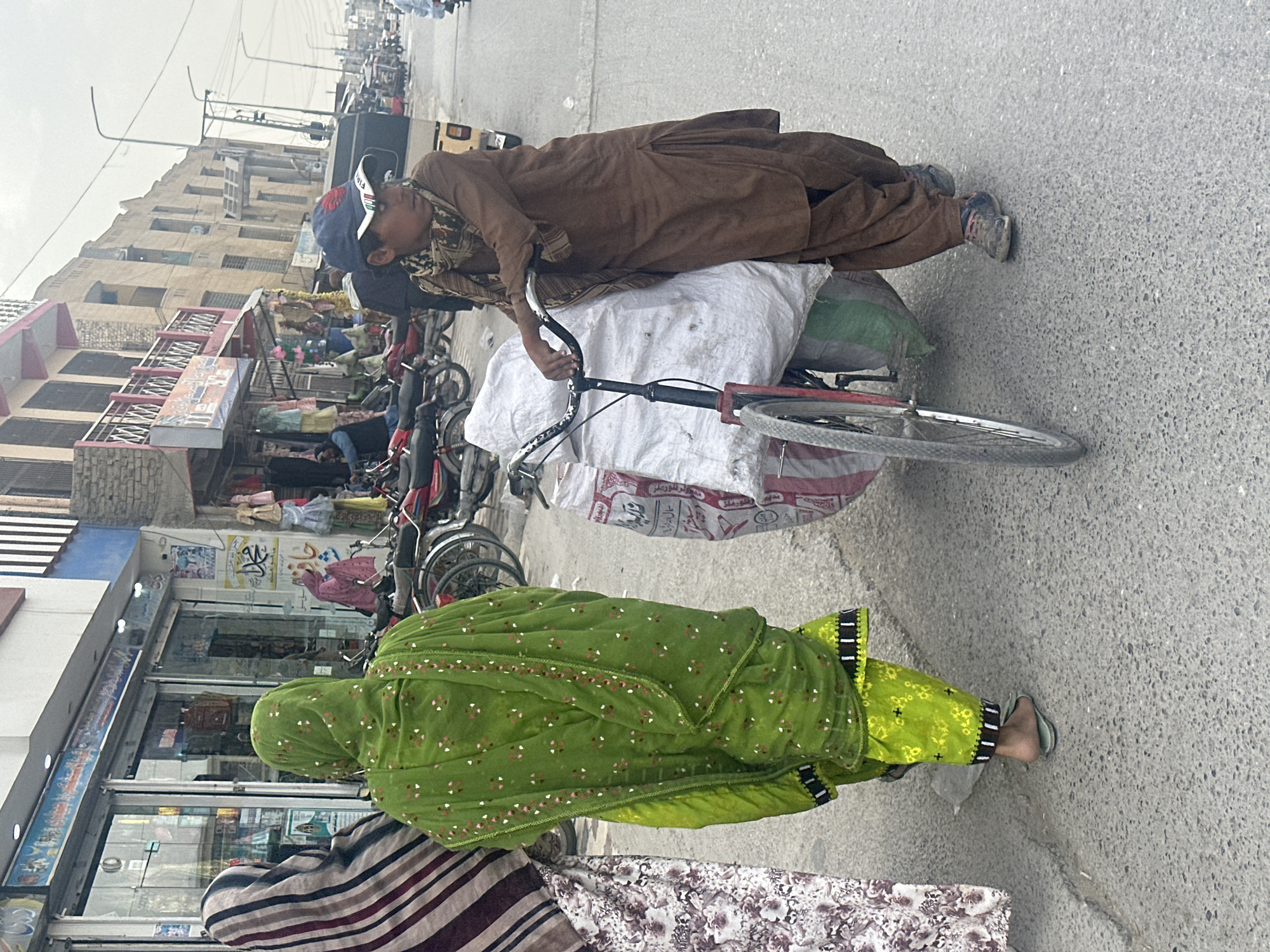 A boy in brown clothing and a cap is leaning over a bicycle loaded with large sacks in Quetta City Tehsil, Pakistan. A woman in green attire walks nearby. Shops line the street in the background with other bicycles and motorbikes parked along the curb.