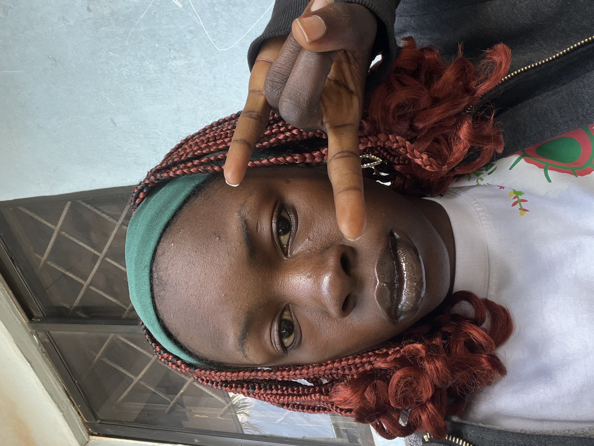 A medium shot captures a person with dark skin, wearing a dark grey zip-up jacket over a white t-shirt adorned with a green and red floral pattern. A green headband is worn across their forehead, and their hair is styled in numerous red braids, some of which are curled at the ends. The individual is looking directly at the camera with their eyes open. Their lips are glossy and appear to be dark brown. The person's right hand is raised, with their index and middle fingers extended outwards in a peace sign gesture, while their thumb and ring finger are curled inwards. The background consists of a light blue wall on the right and a window with a metal grate on the left. The image appears to have been taken outdoors, potentially in the city of Jos, Nigeria, given the architecture and lighting.