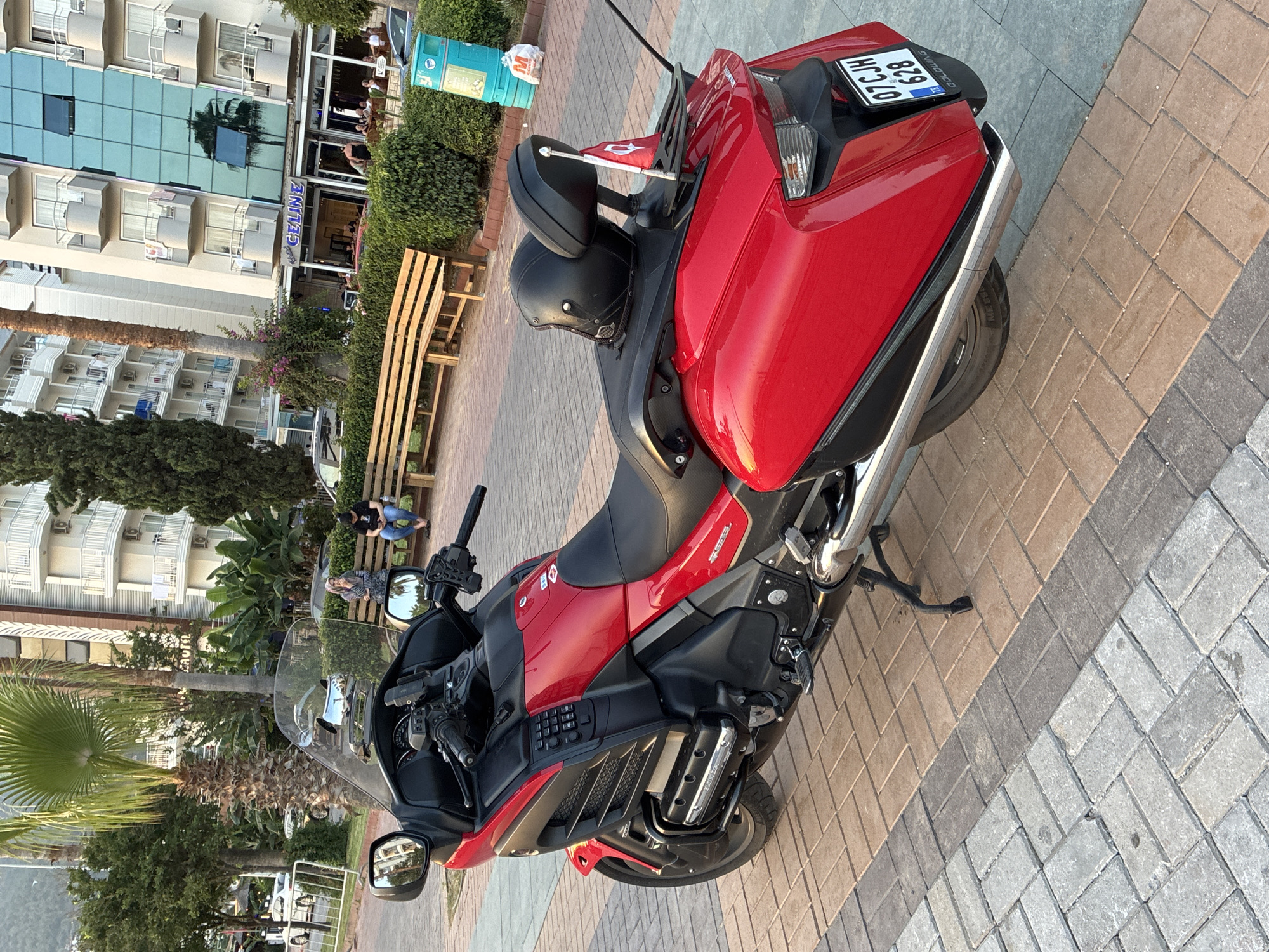 A bright red Honda motorcycle is parked on a pedestrian walkway composed of interlocking bricks of various colors. The motorcycle has a large windshield, black leather seats, and a black helmet resting on the rear seat. A small Turkish flag is attached to the back of the motorcycle. In the background, a multi-story building with balconies and a glass facade stands. Several palm trees and other greenery are visible, suggesting a warm climate. The building's signage reads "CELINE". Two individuals are seated on a wooden bench to the left of the motorcycle. A blue trash bin is located near the bench. The scene appears to be in a public area within the city of Alanya, Turkey, likely a promenade or street.